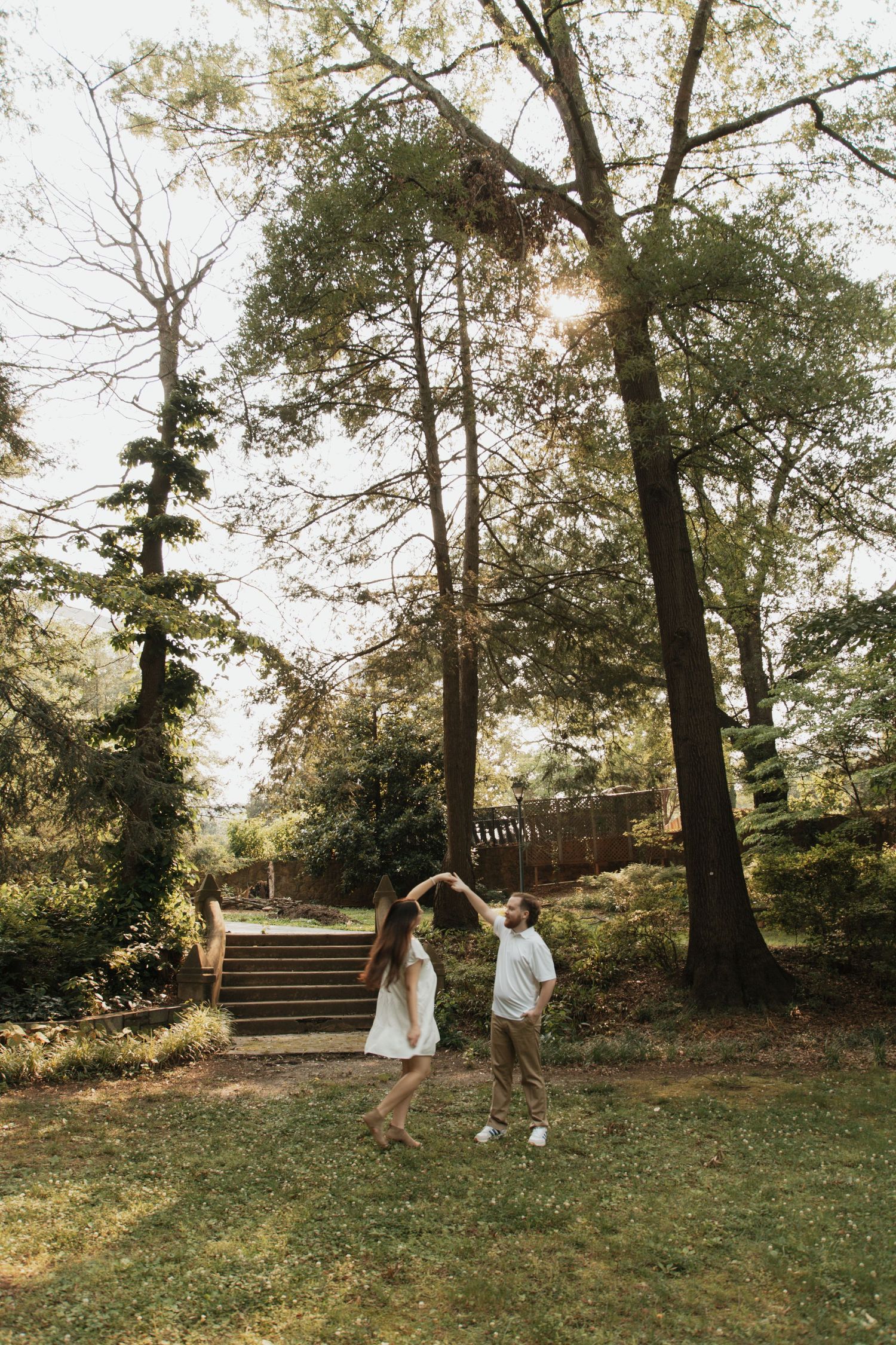Couple dancing together in a lush green park, surrounded by tall trees and wooden steps in warm sunlight.