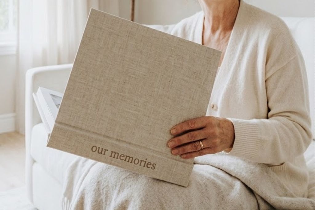 Elderly woman holding a linen-covered photo album labeled 'our memories' while seated on a white sofa.