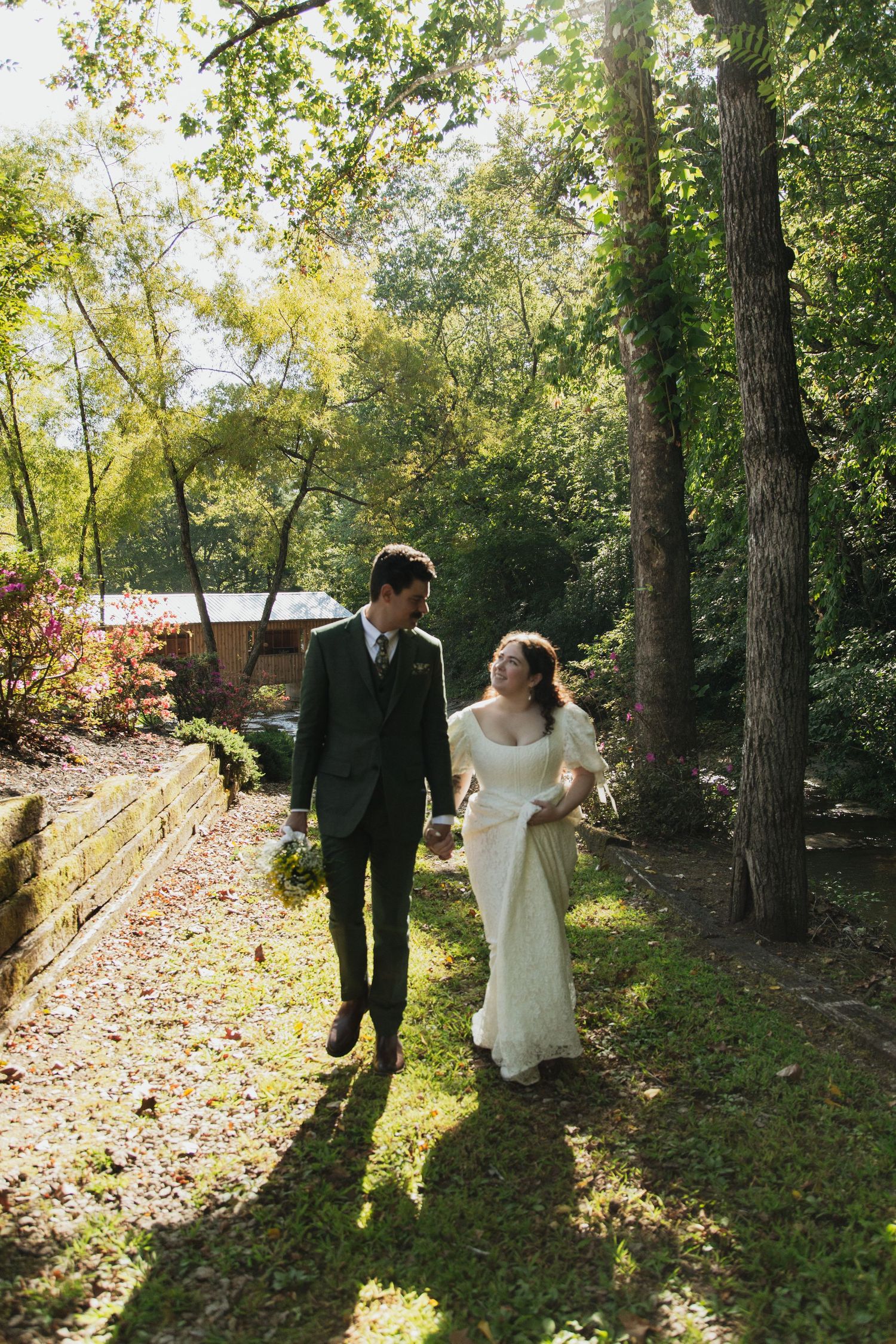 Bride and groom walk hand-in-hand through a sunlit garden path surrounded by lush green trees.  venue at ballenger bridge landrum sc