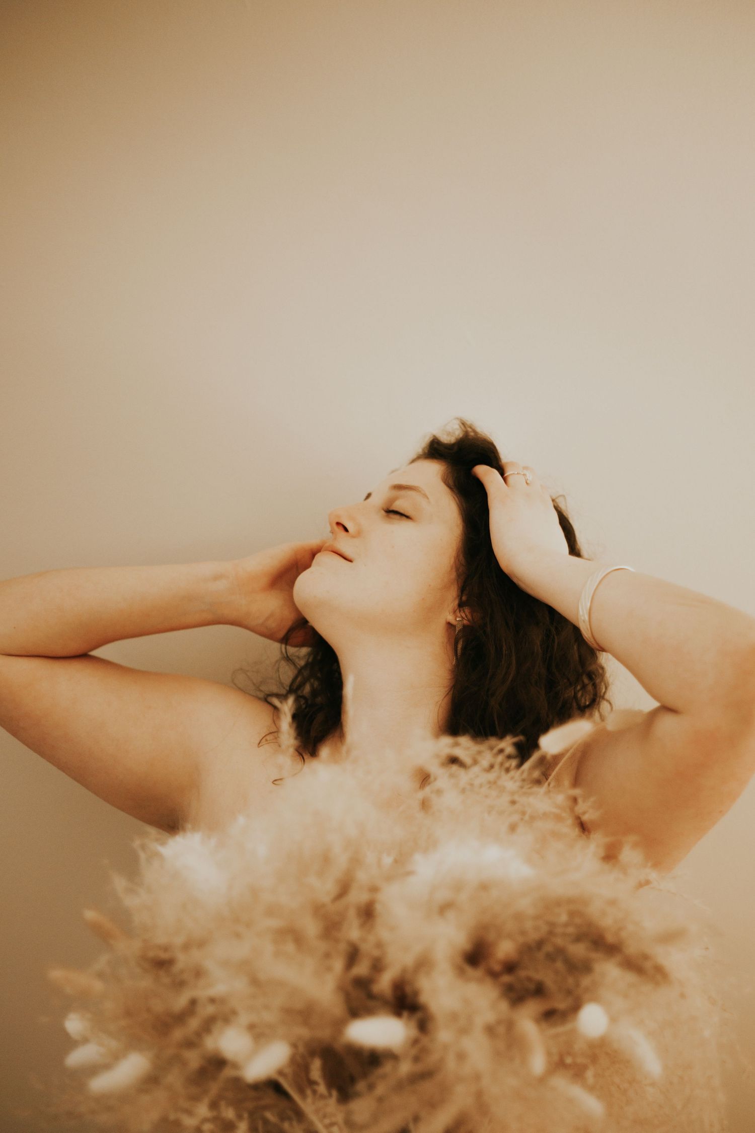 A woman with dark hair poses with eyes closed, hands in hair, holding dried pampas grass bouquet.