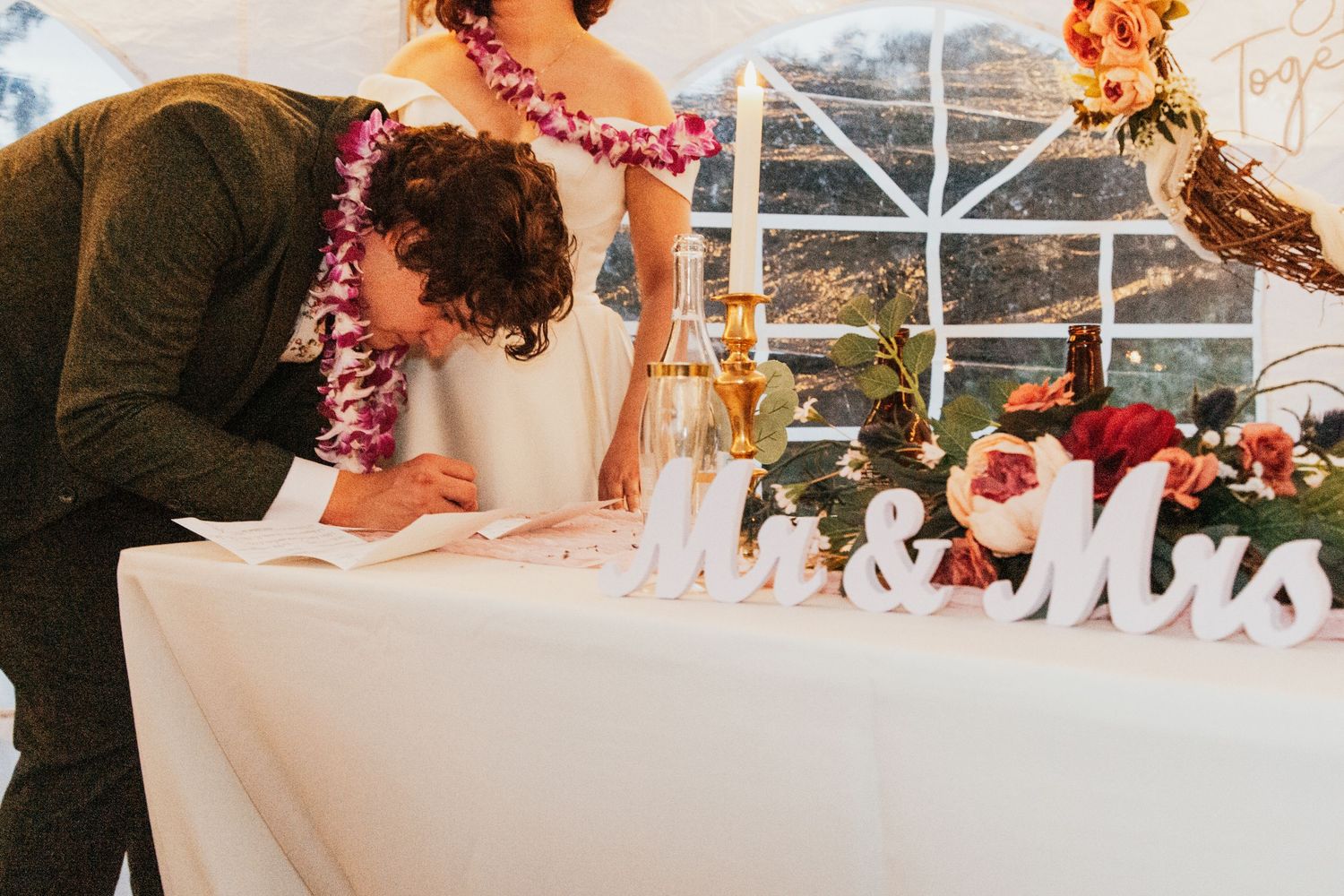 Bride and groom signing wedding registry at decorated table with Mr & Mrs letters and floral arrangements.