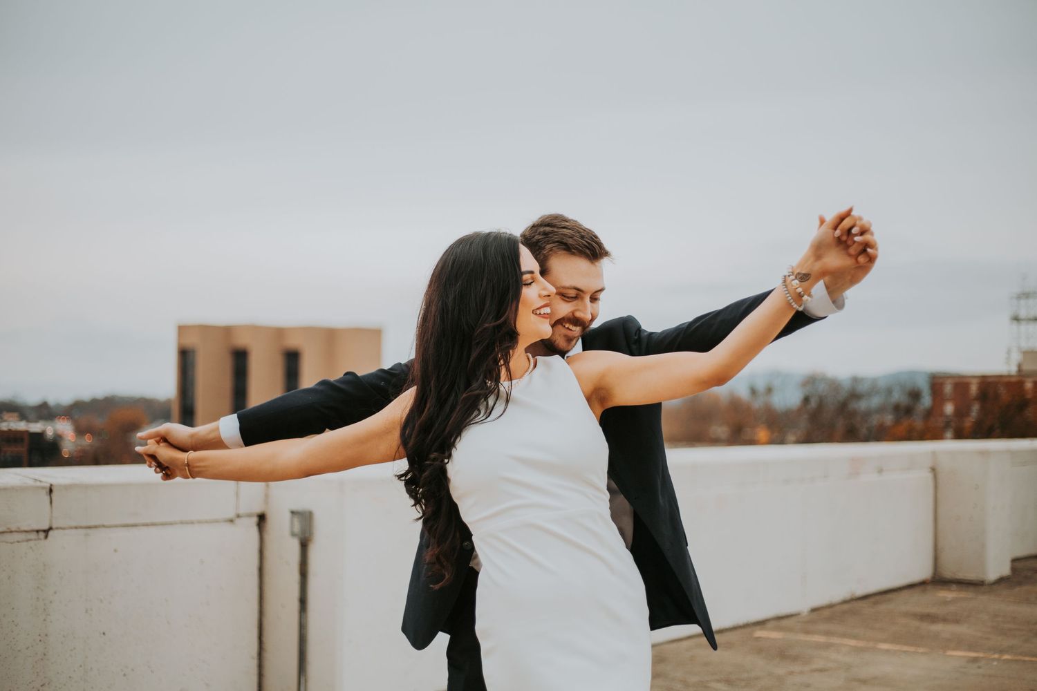 Happy couple dancing on rooftop, woman in white dress and man in suit, arms outstretched, celebrating together.