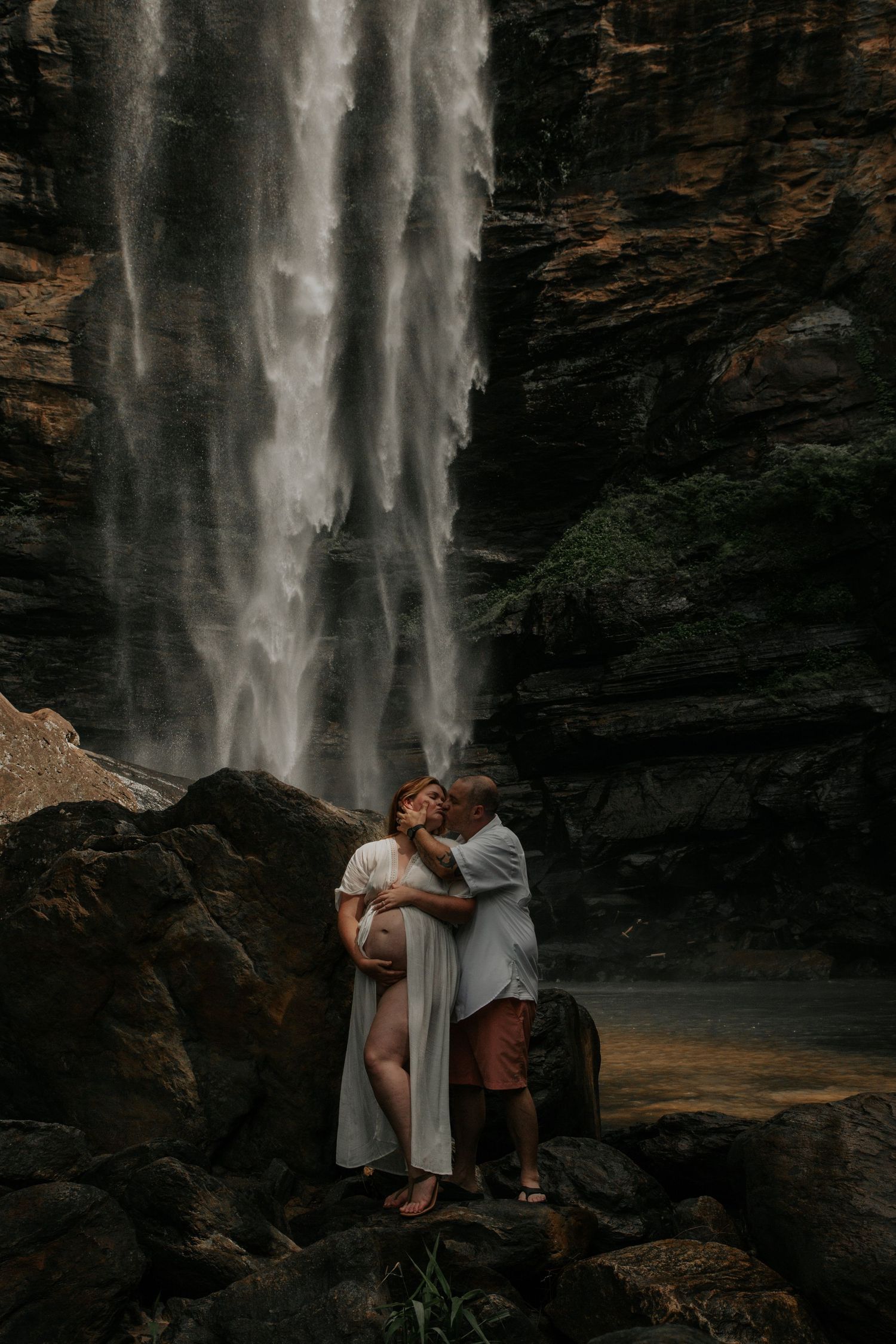 Expecting couple sharing a tender kiss in front of a stunning cascading waterfall on rocky terrain. toccoa falls toccoa georgia