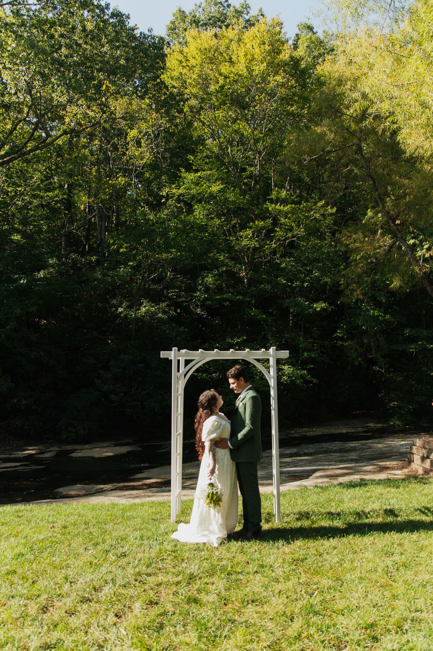 Bride and groom standing beneath white wooden arch arbor on sunny green lawn with lush tree backdrop. venue at ballenger bridge landrum sc