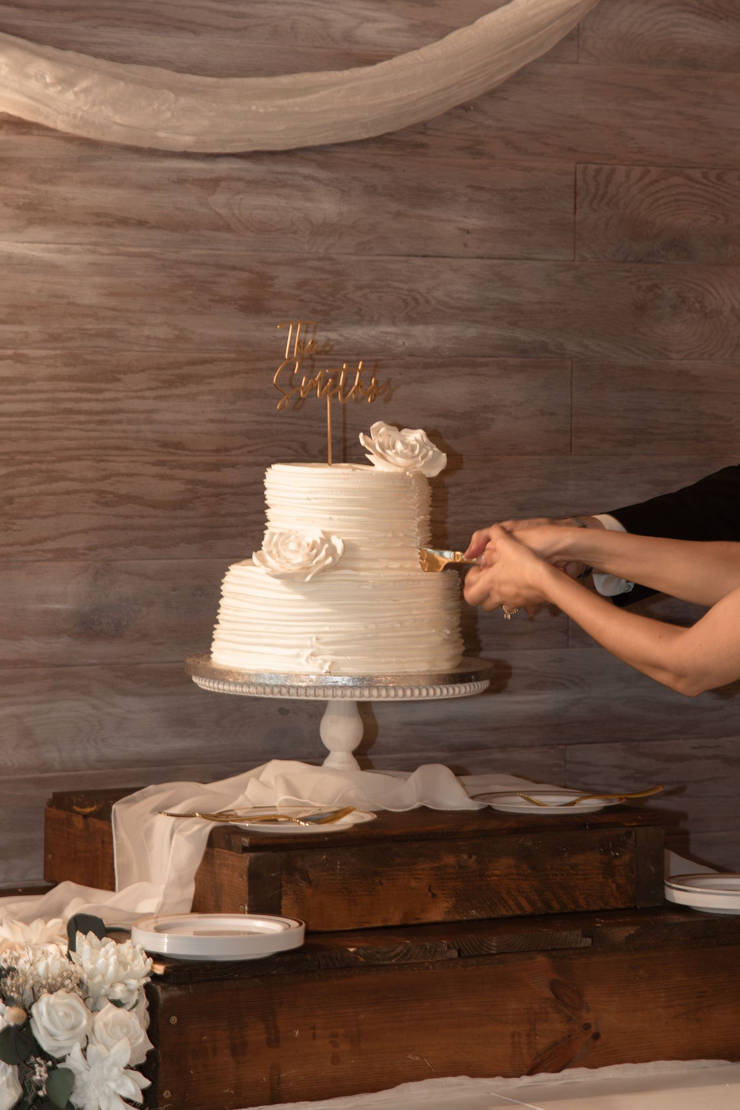 Two-tier white wedding cake with floral decorations and gold topper being cut on wooden stand.