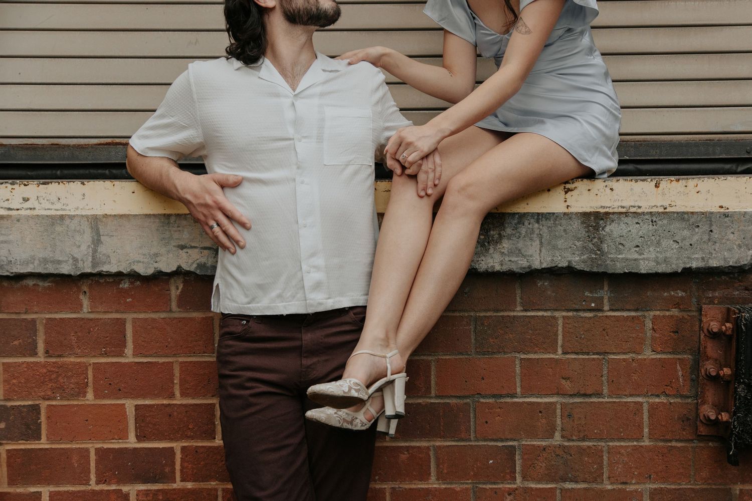 A couple poses romantically against a brick wall, man in white shirt and brown pants, woman in gray dress with heels.