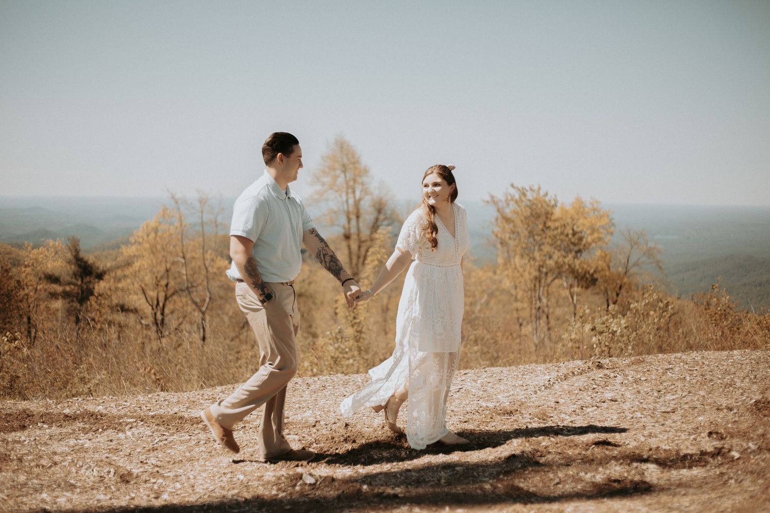 Couple holding hands and dancing on a hilltop in autumn, woman in white dress, man in casual gray outfit.