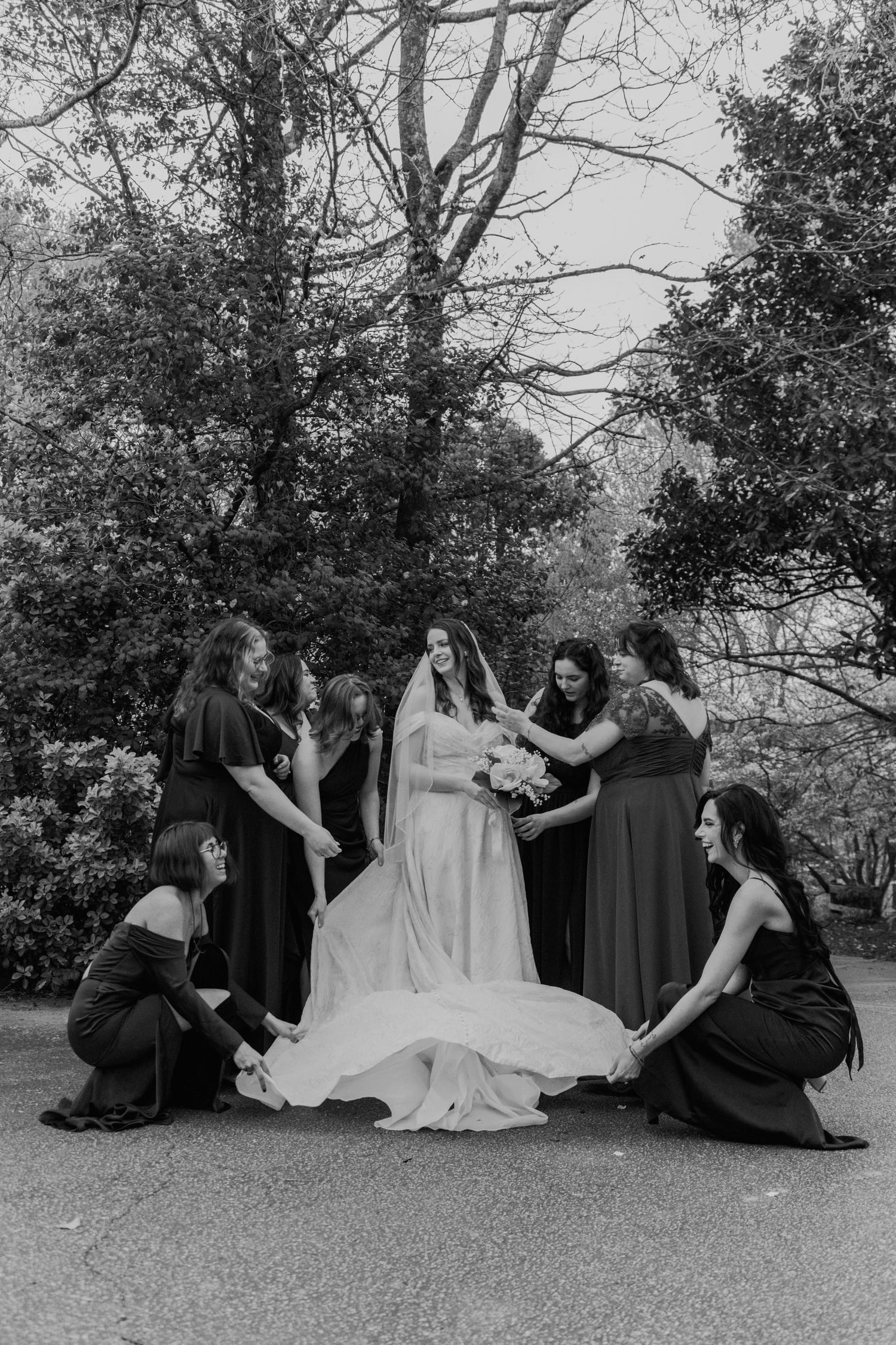 Black and white photo of a bride surrounded by bridesmaids in dark dresses adjusting her gown and veil outdoors.