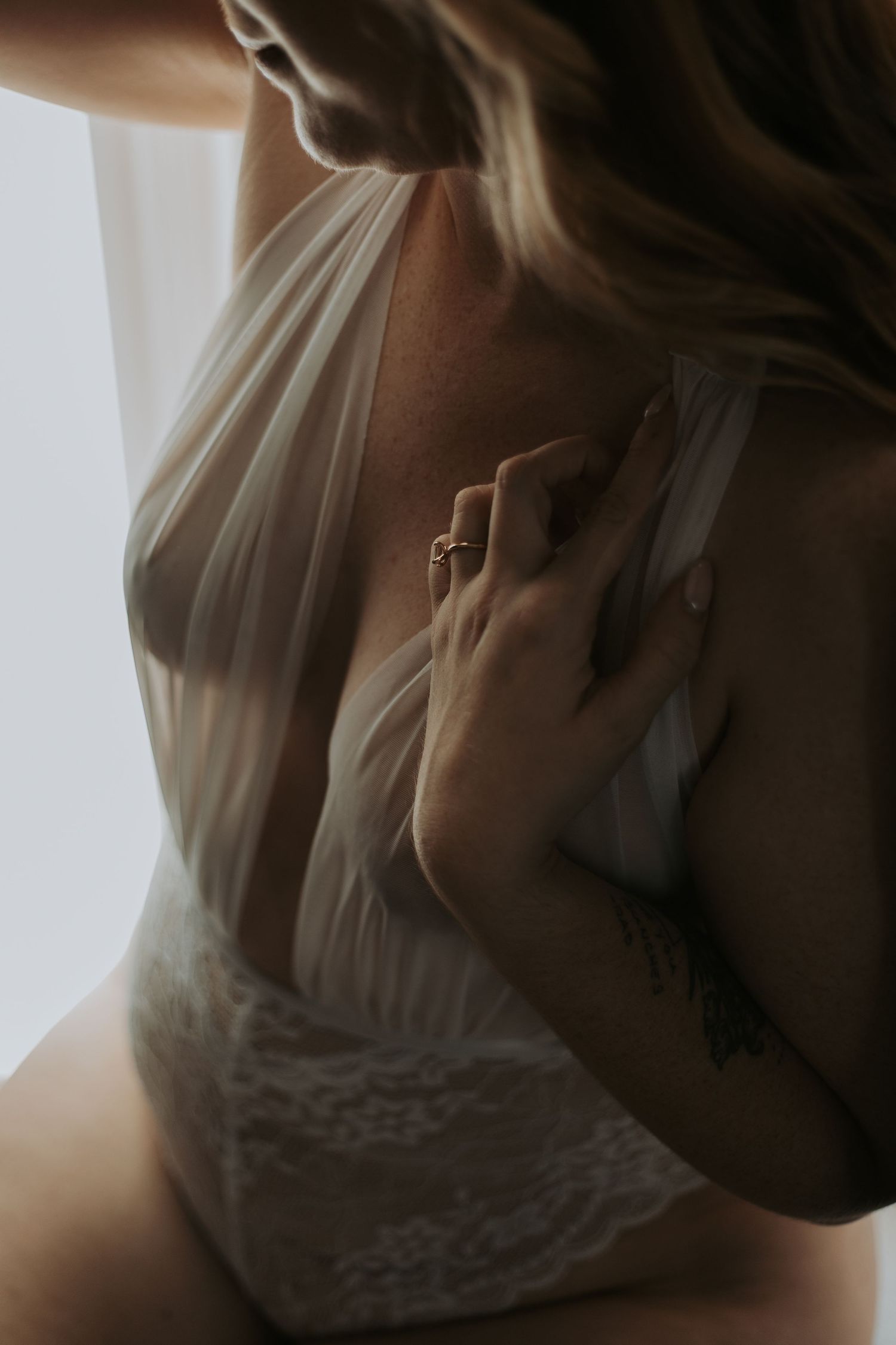 Close-up of woman in sheer white lace bodysuit with ring and tattoo in soft boudoir lighting.. wanderlust boudoir studio honea path sc