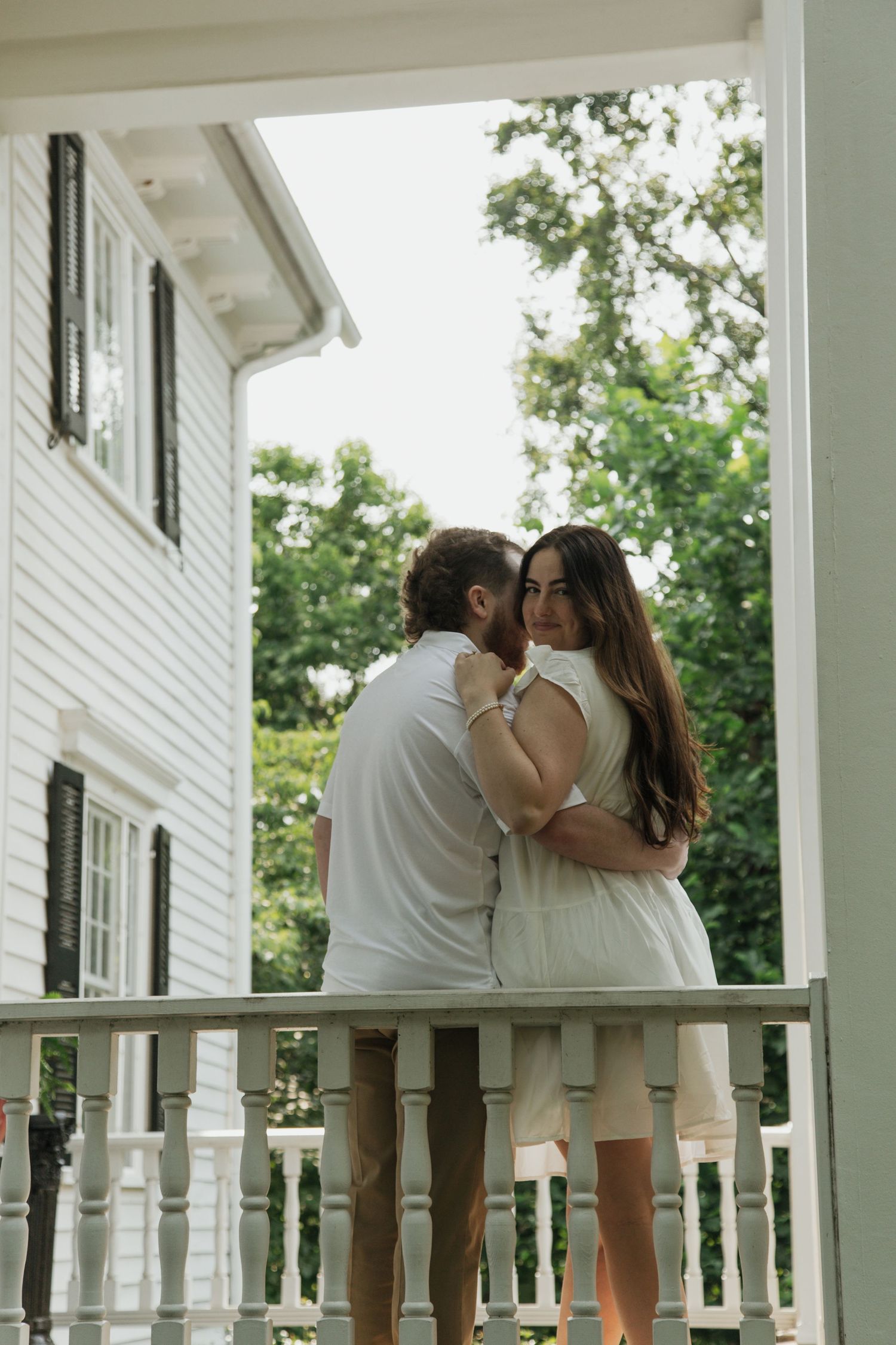 Couple embracing tenderly on white porch balcony with lush green trees in background. kilgore lewis house and garden greenville sc