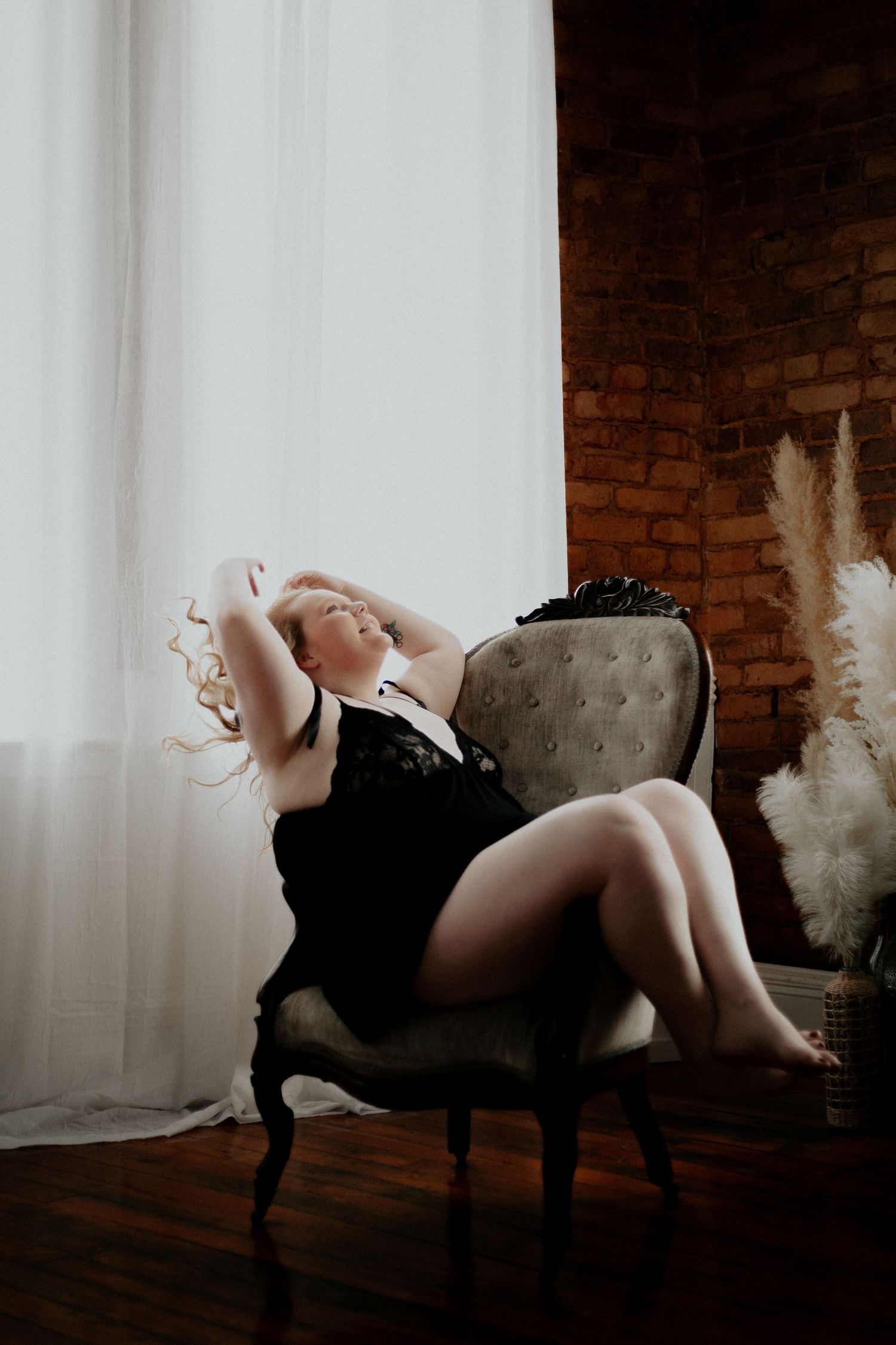 Blonde woman in black lace teddy reclining in vintage chair by window in boudoir photography session.. wanderlust boudoir studio honea path
