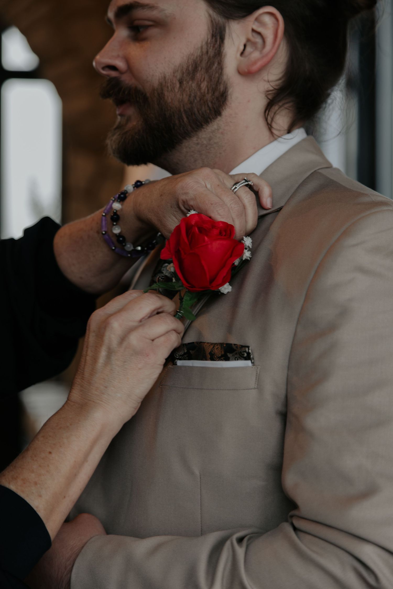 A bearded groom in a tan suit has a red rose boutonniere pinned to his lapel.