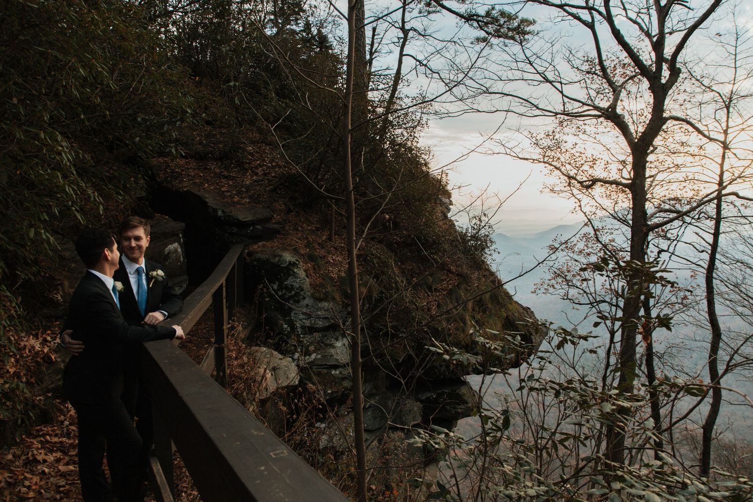 Two grooms in suits stand on a scenic overlook surrounded by autumn trees and misty mountain views at their outdoor wedding.
