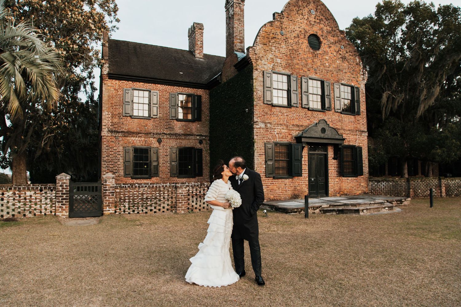 Bride and groom share romantic kiss in front of historic red brick plantation house at outdoor wedding venue.