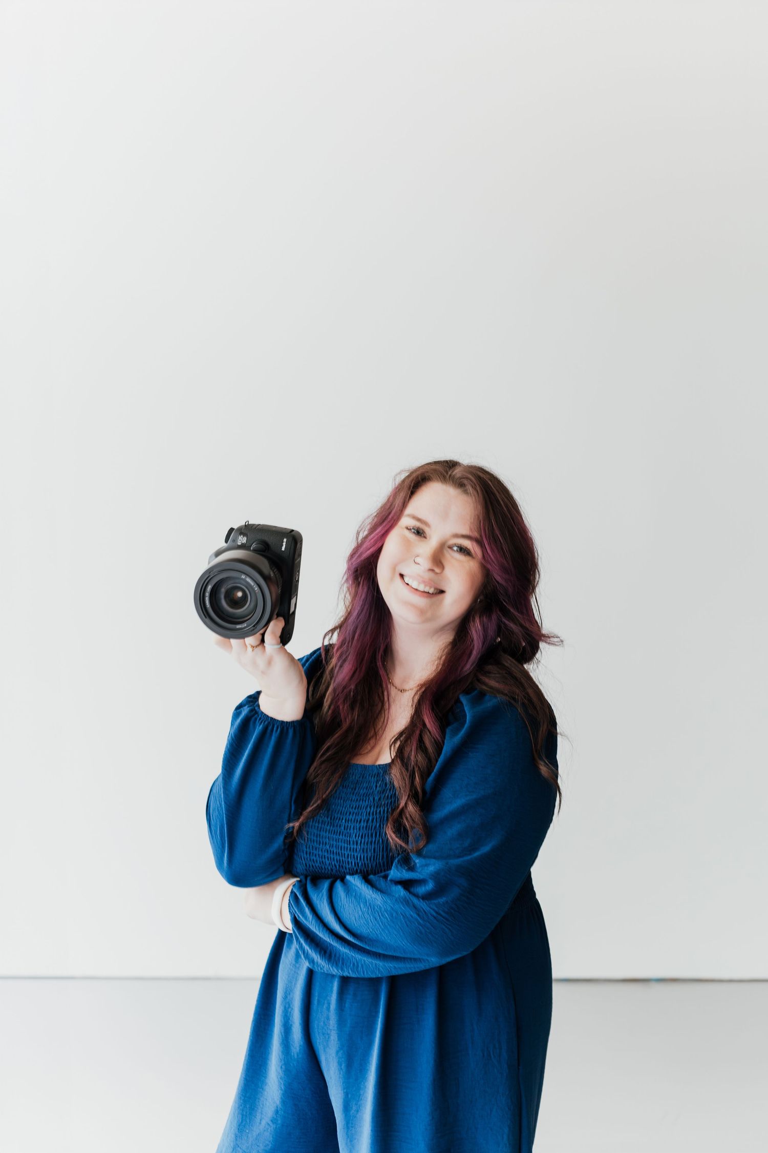 Smiling woman with long hair holding a DSLR camera, wearing a blue dress against a white background.