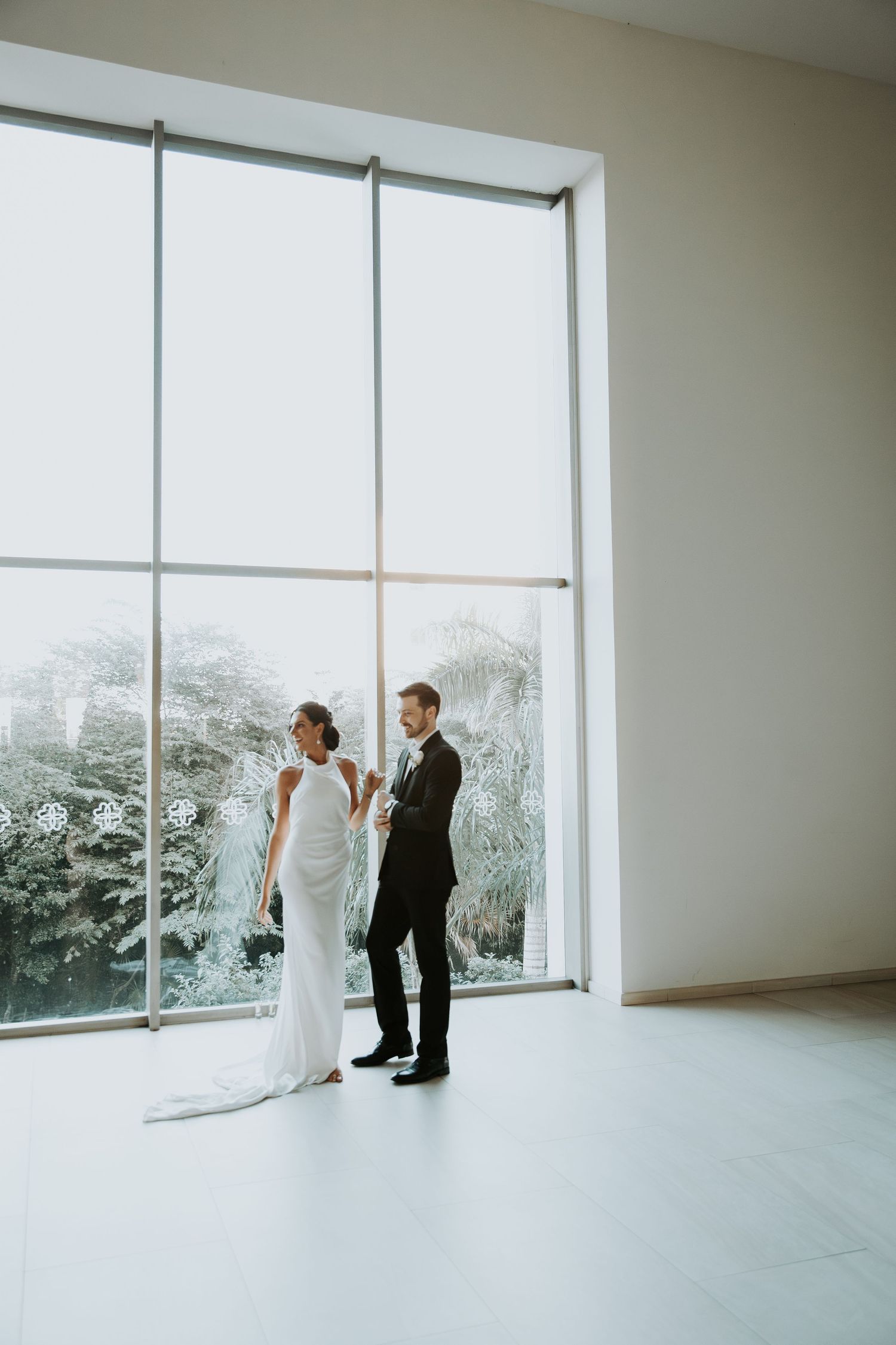 Bride in white gown and groom in black suit stand before large floor-to-ceiling windows in elegant venue.