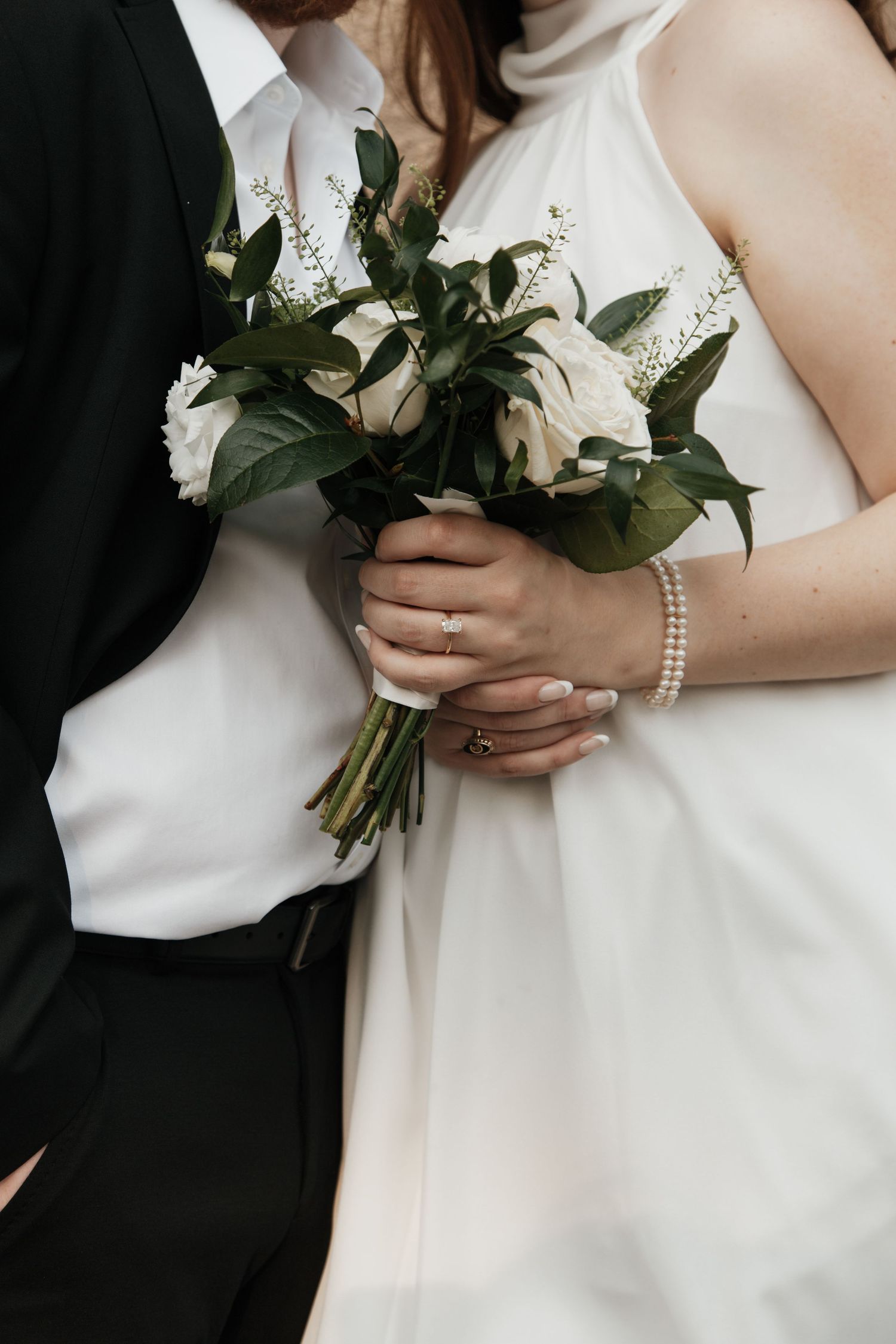 Bride in white dress holding elegant bouquet of white roses and greenery with groom in black suit.