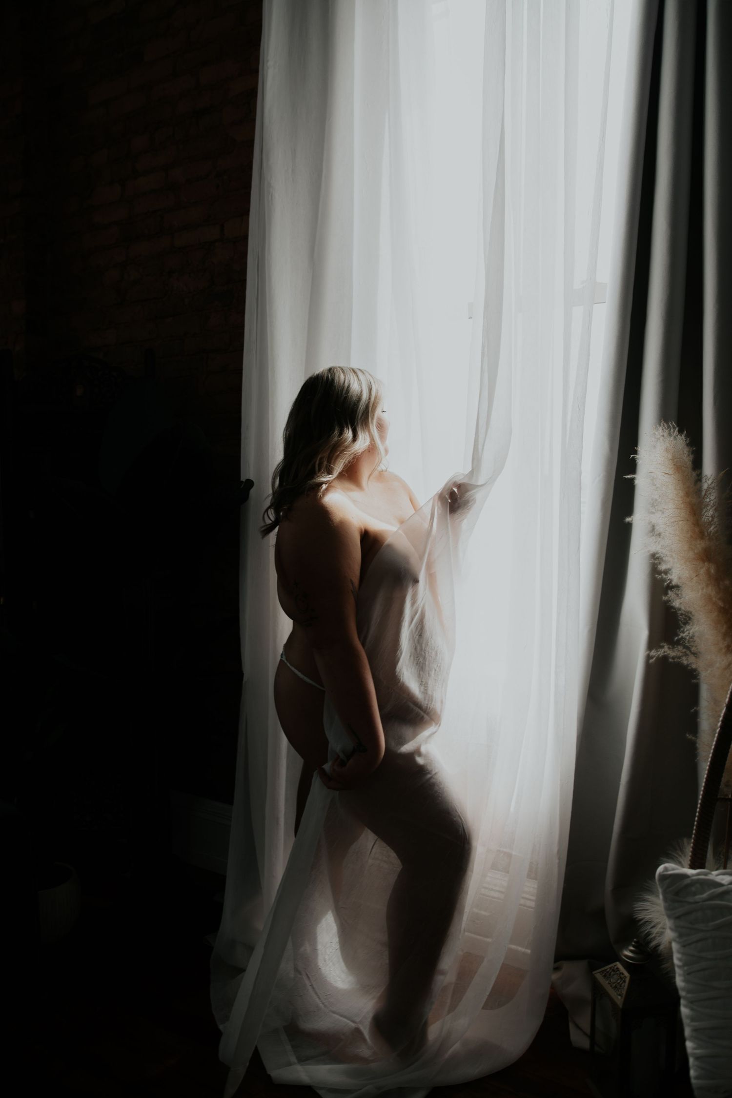 Silhouette of woman in white lingerie standing by sheer curtains in moody boudoir photography.