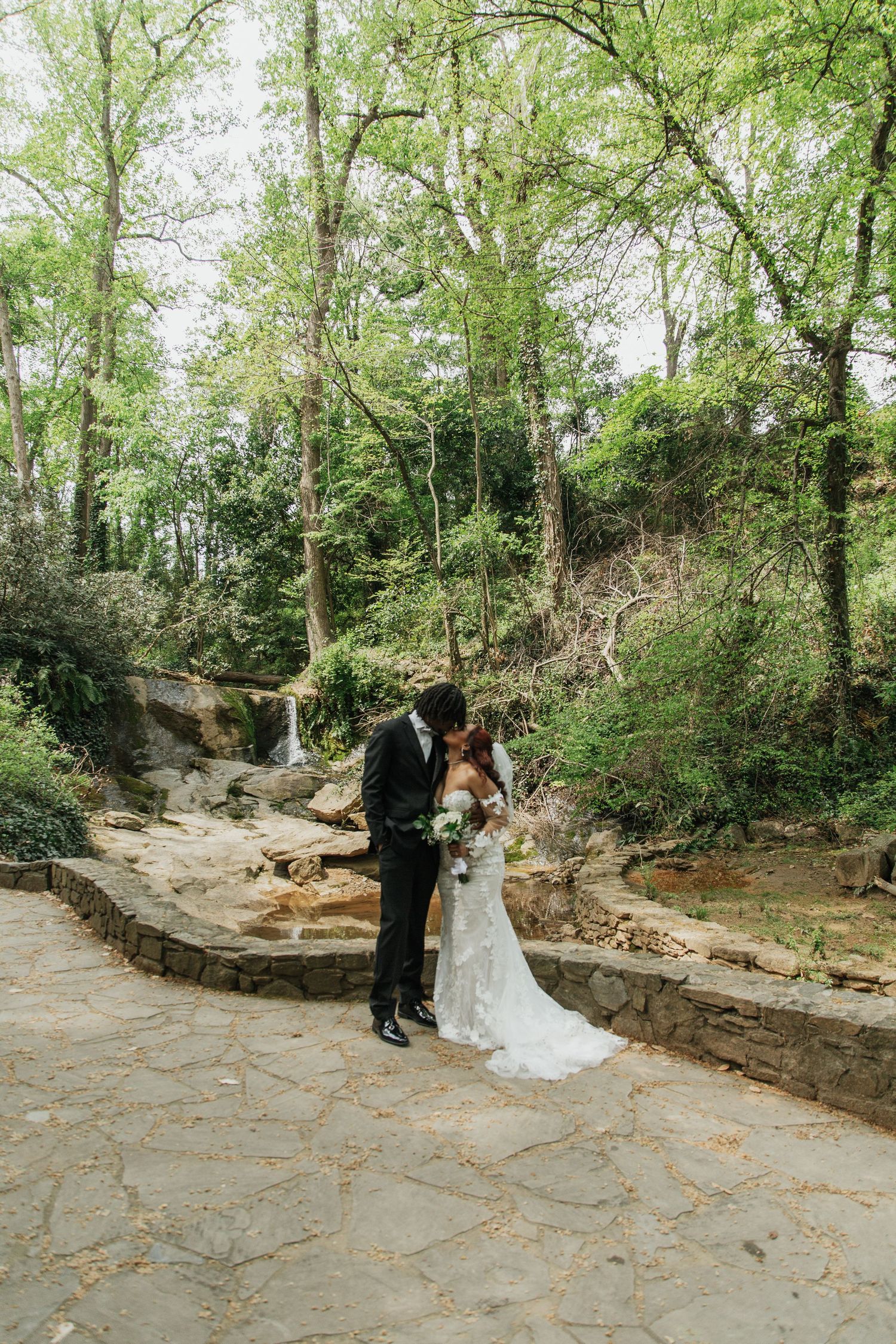 Bride and groom share a romantic kiss on a stone path surrounded by lush greenery and a waterfall backdrop.