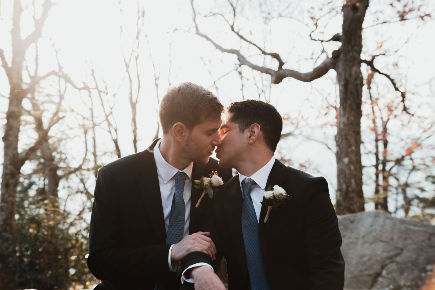 Two grooms in dark suits share a kiss outdoors among bare trees on their wedding day.
