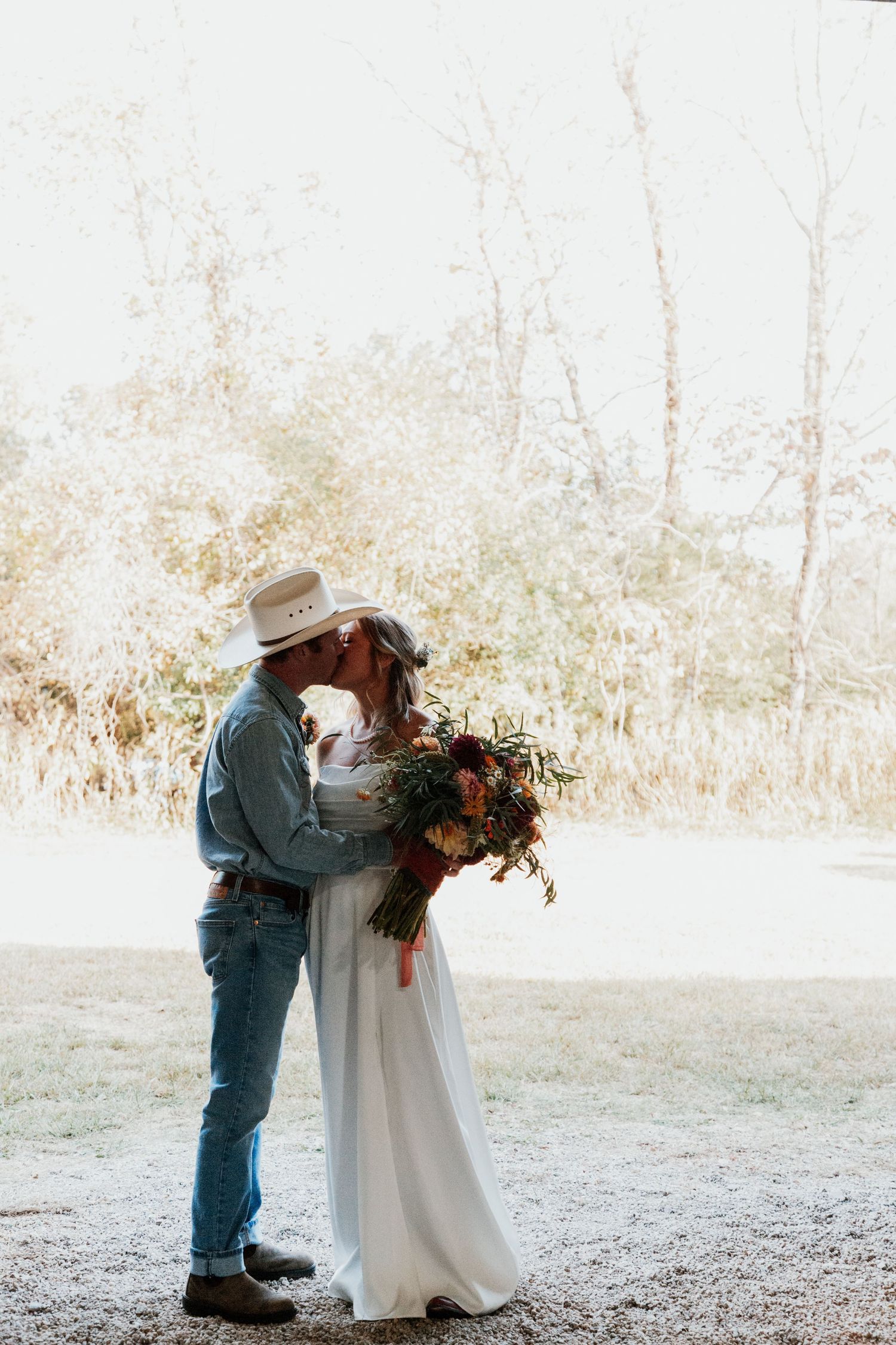 Couple in cowboy hats sharing a kiss at outdoor wedding, bride in white gown holding colorful bouquet.