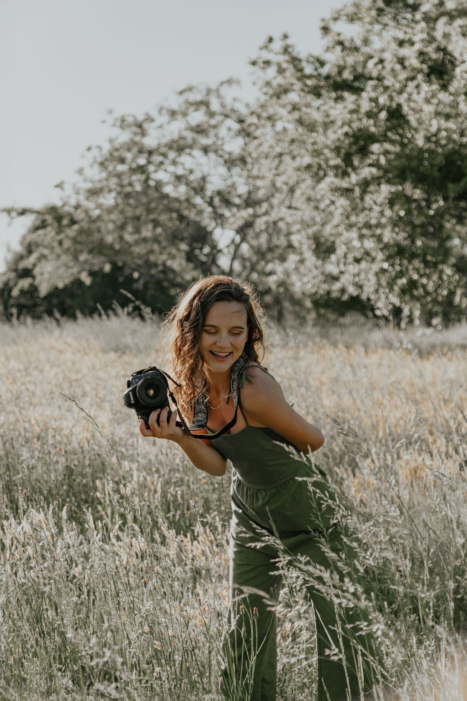 A smiling woman in a green jumpsuit holds a camera in a sunlit field with tall grass and blooming trees.