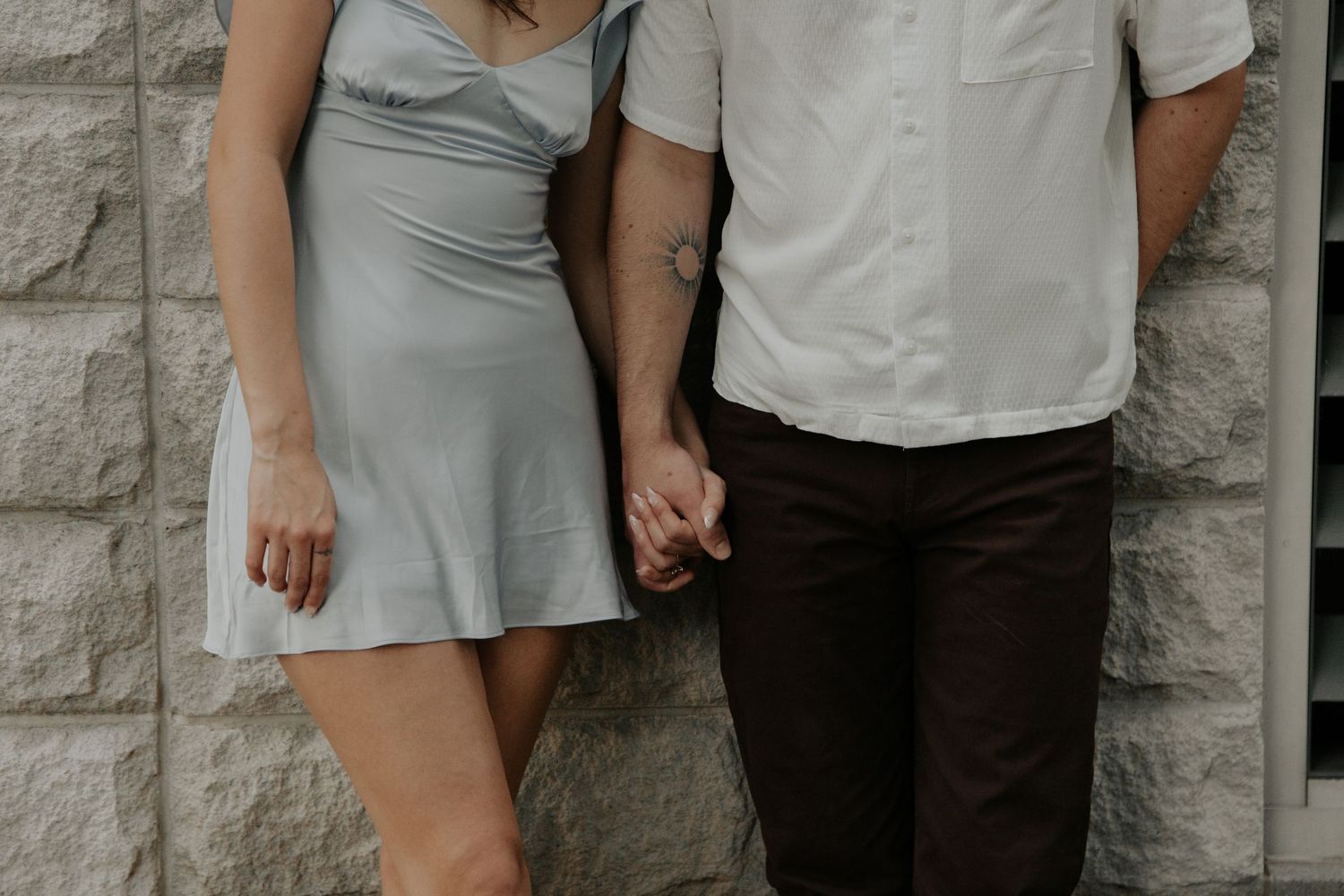 Couple holding hands in front of stone wall, woman in silver mini dress, man in white shirt and dark pants.