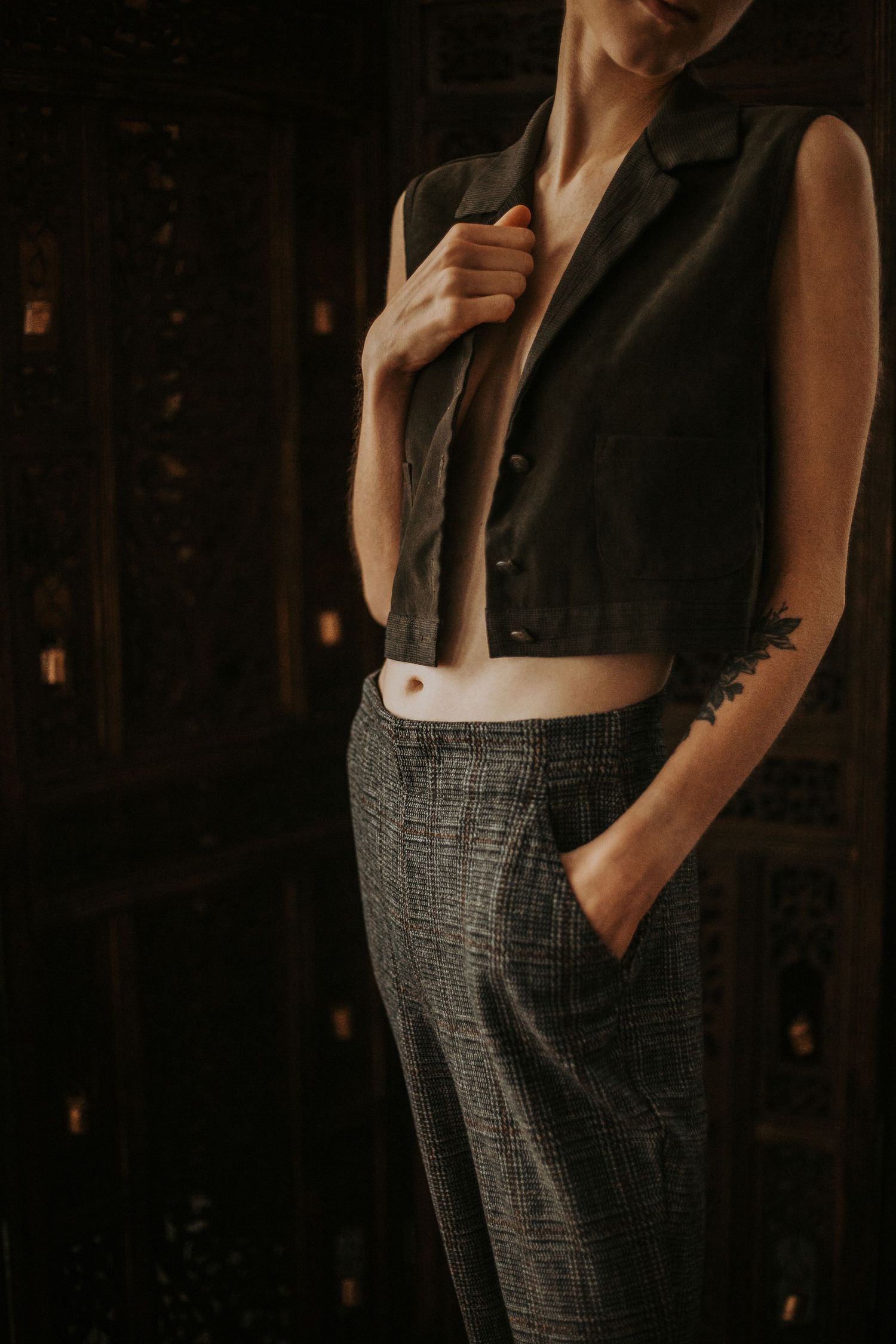 A slender figure in a cropped vest and plaid trousers reveals a floral arm tattoo in moody dark lighting.