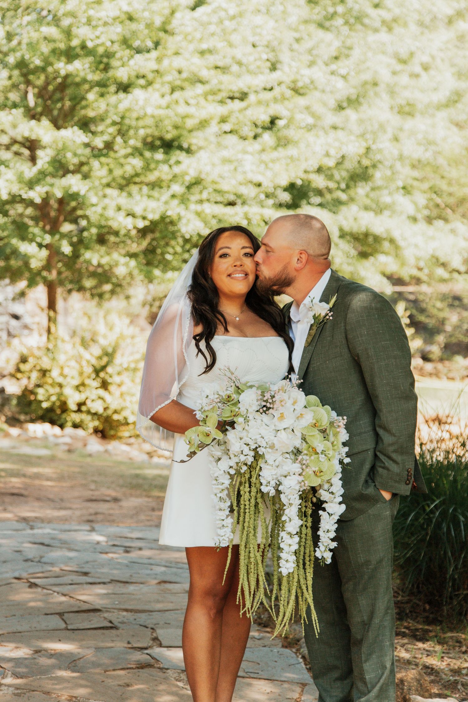 Bride in white mini dress with cascading floral bouquet receives kiss from groom in green suit outdoors.