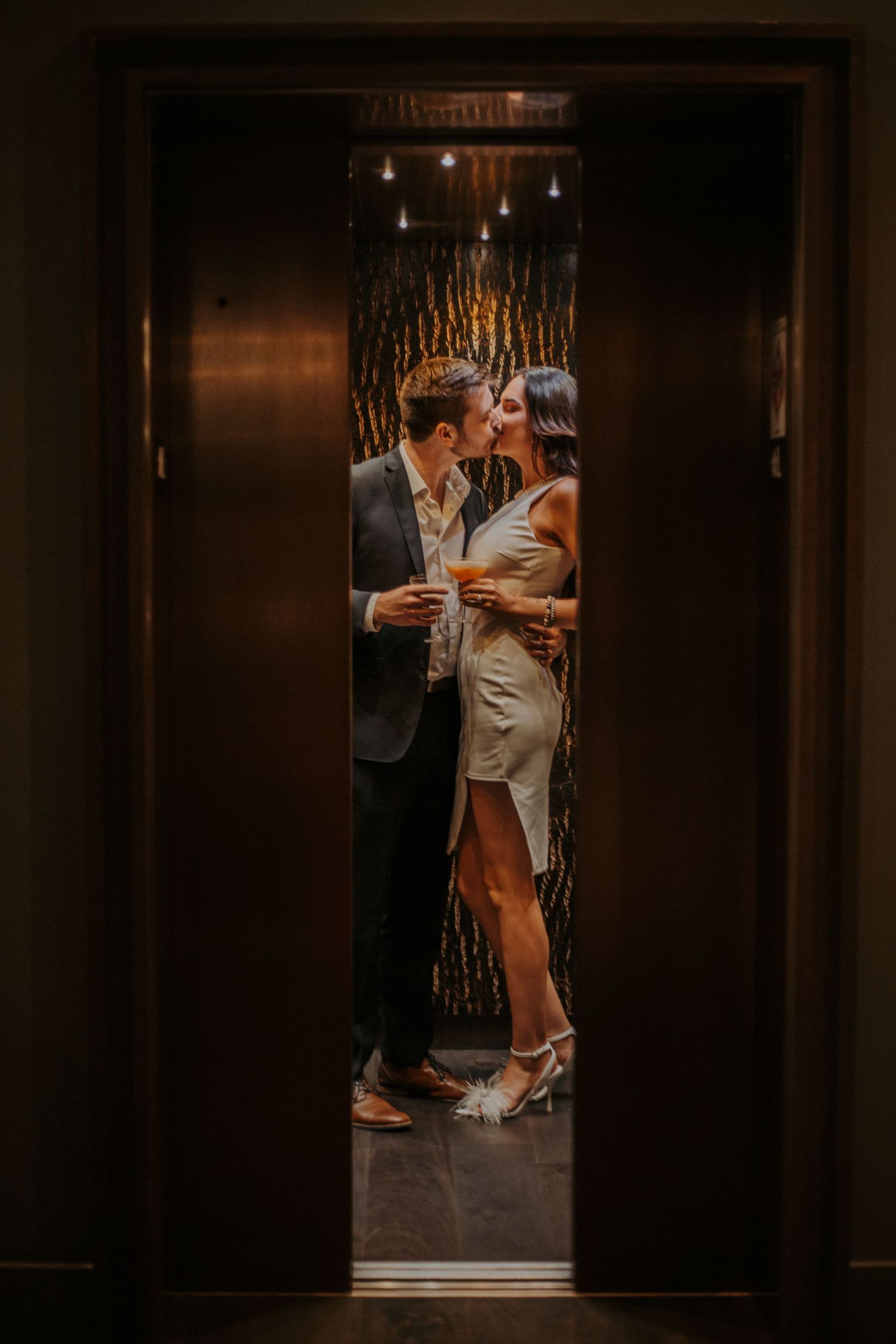 Couple sharing a romantic kiss in a warmly lit elevator, dressed in elegant evening attire.