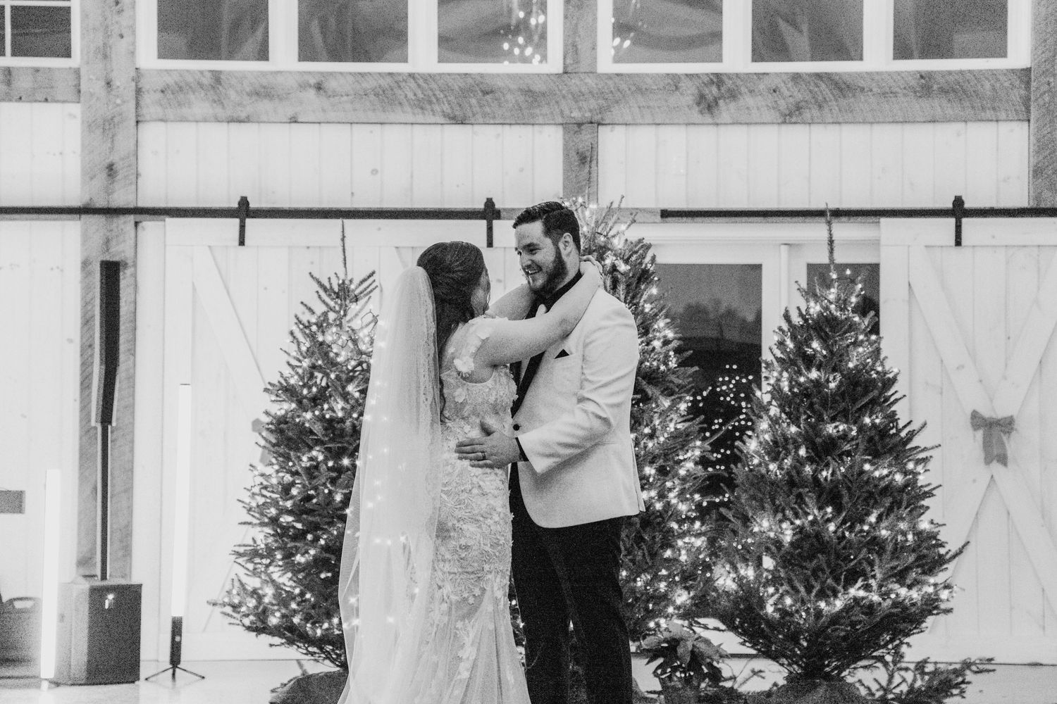 Black and white photo of newlyweds sharing a tender moment in front of decorated Christmas trees inside a barn venue. heyward manor (iva, sc