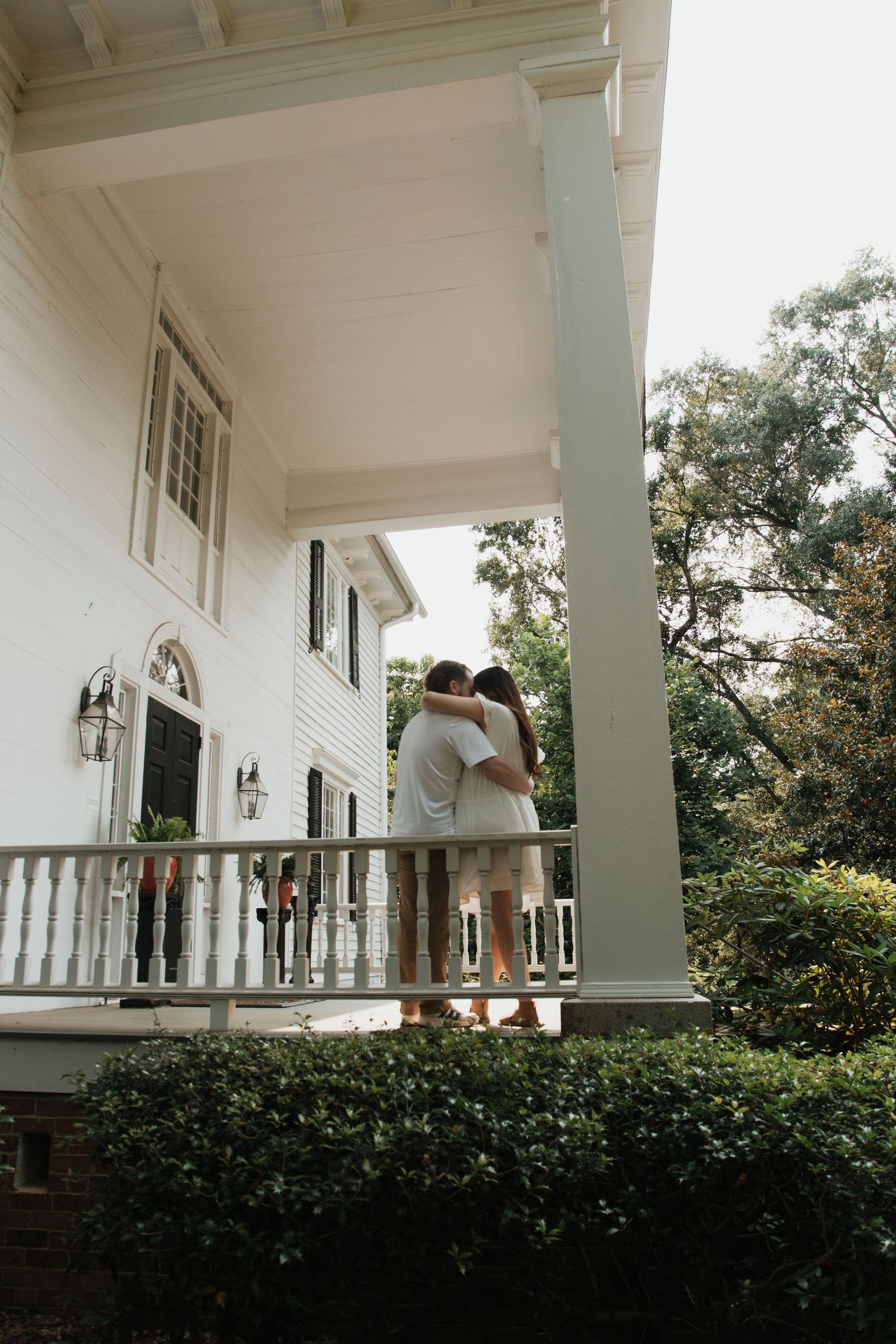 Couple embracing on a white Southern mansion porch surrounded by lush greenery and classic columns.