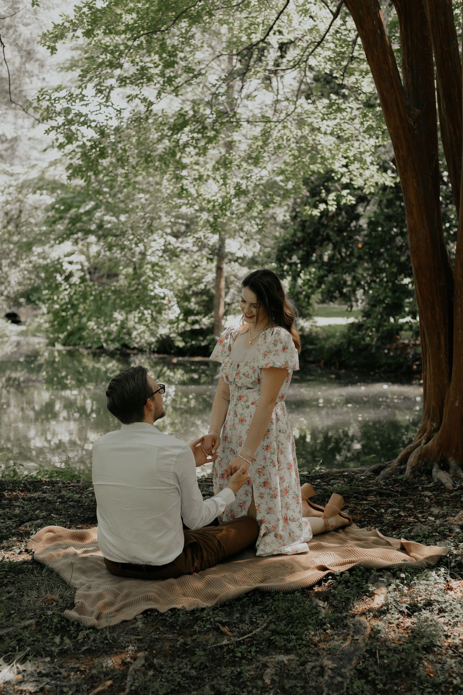 Couple sharing a romantic picnic by a serene pond in a lush green park, woman in floral dress smiling. kilgore lewis house greenville sc