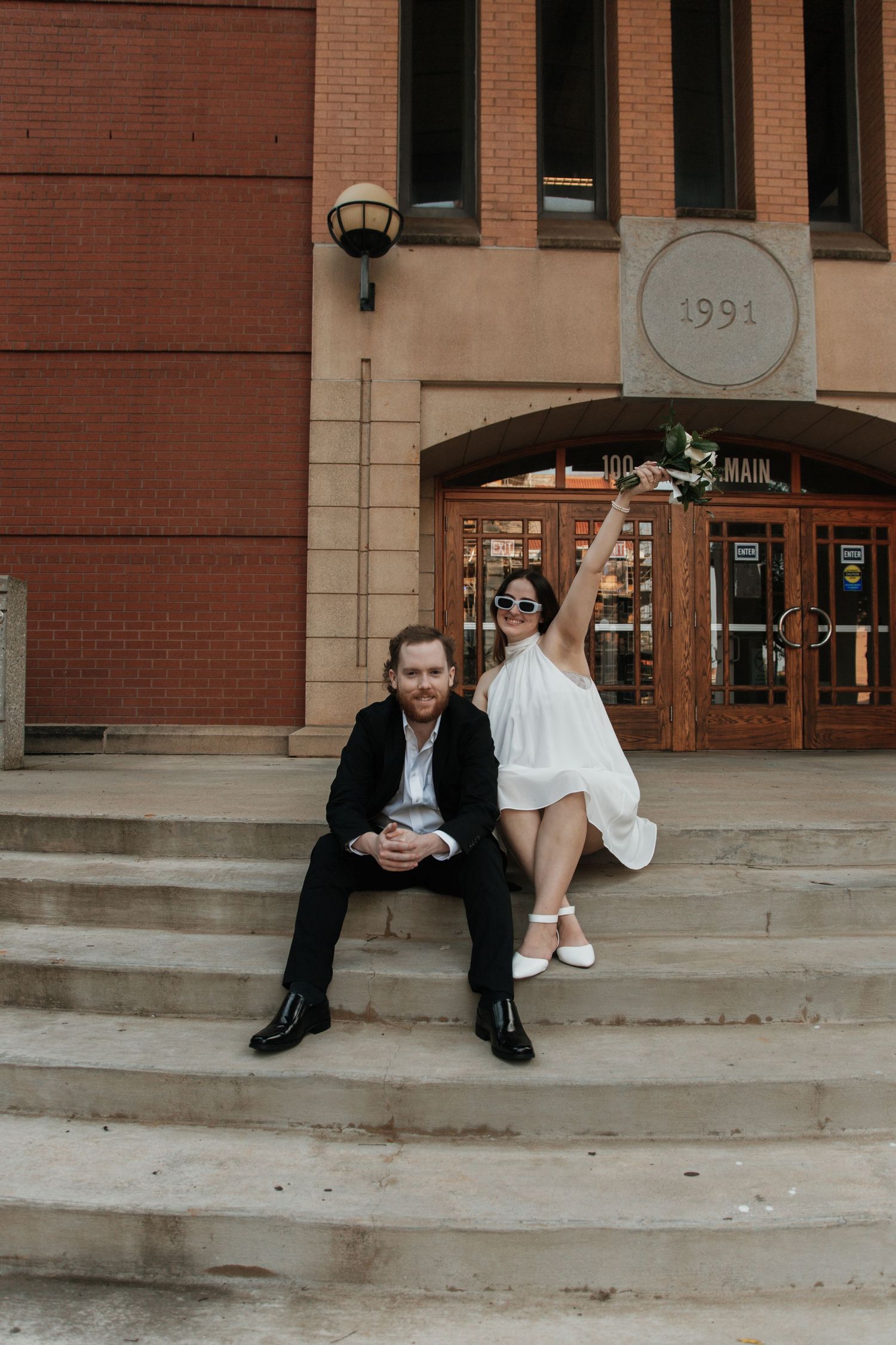 Bride raising bouquet joyfully while groom sits on steps of brick building dated 1991. downtown anderson court house sc