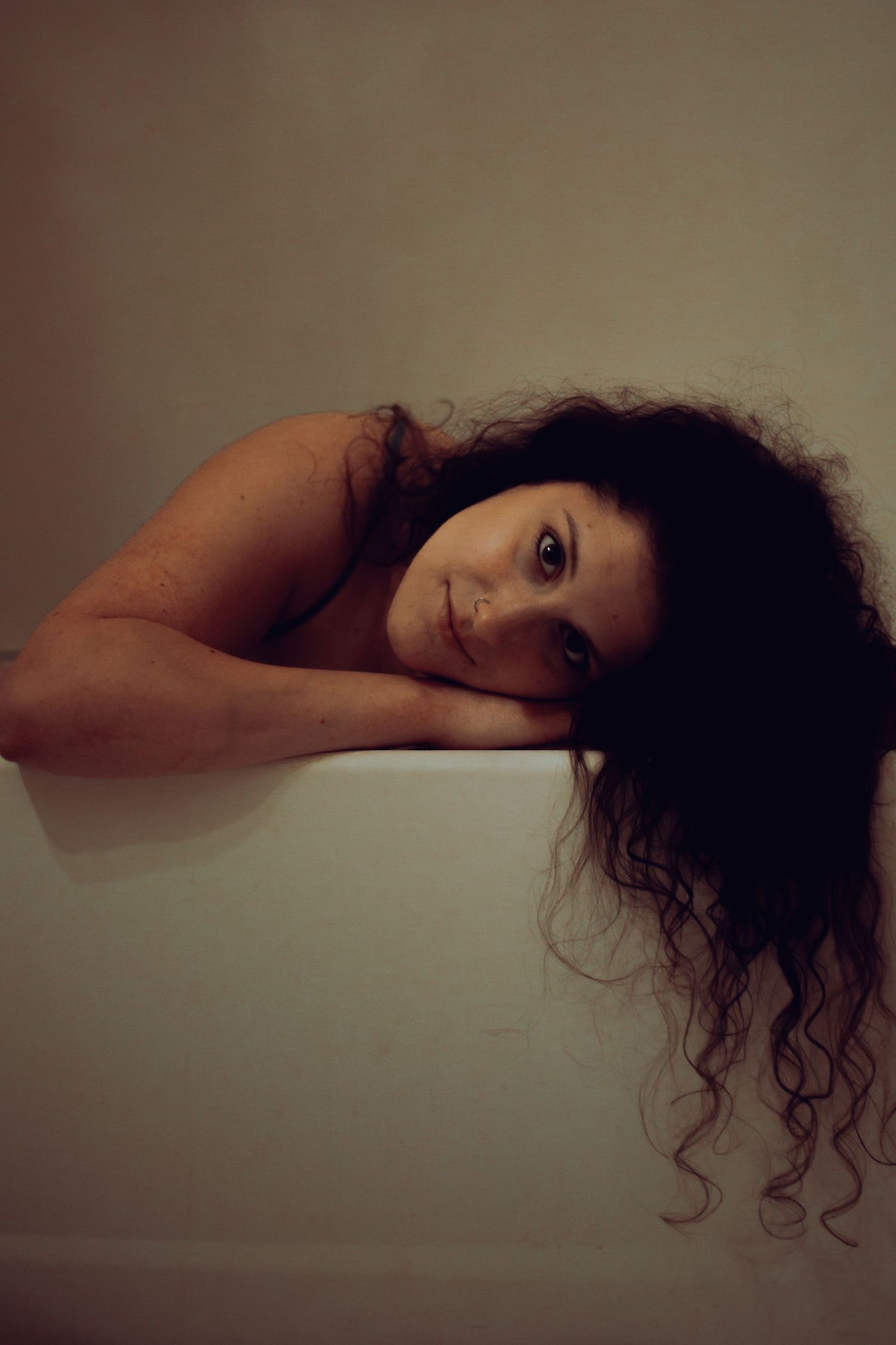 Young woman with curly dark hair resting her chin on arms over a white bathtub edge.