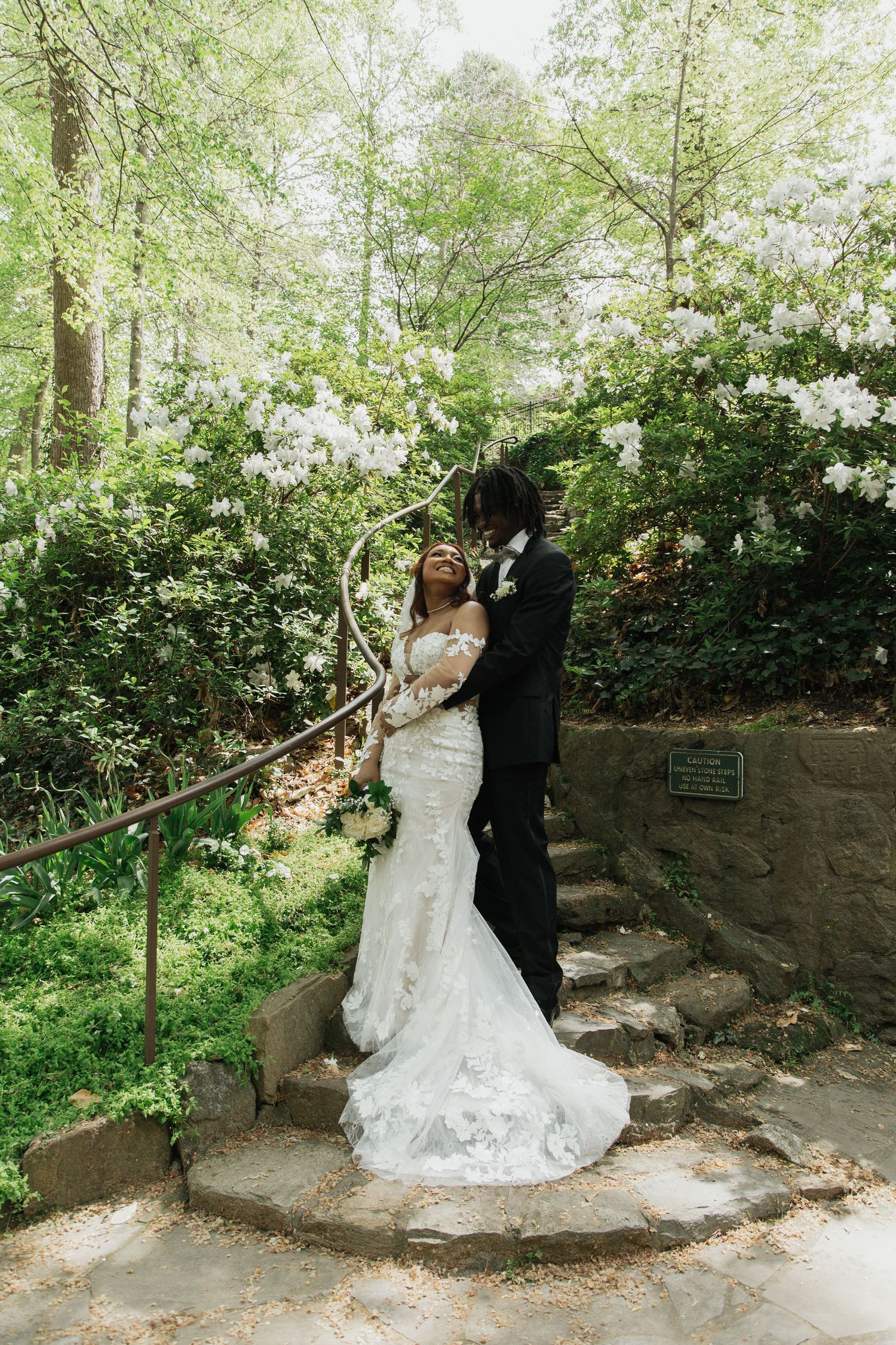 Bride and groom embrace on stone steps surrounded by blooming white flowers in garden. falls park greenville sc