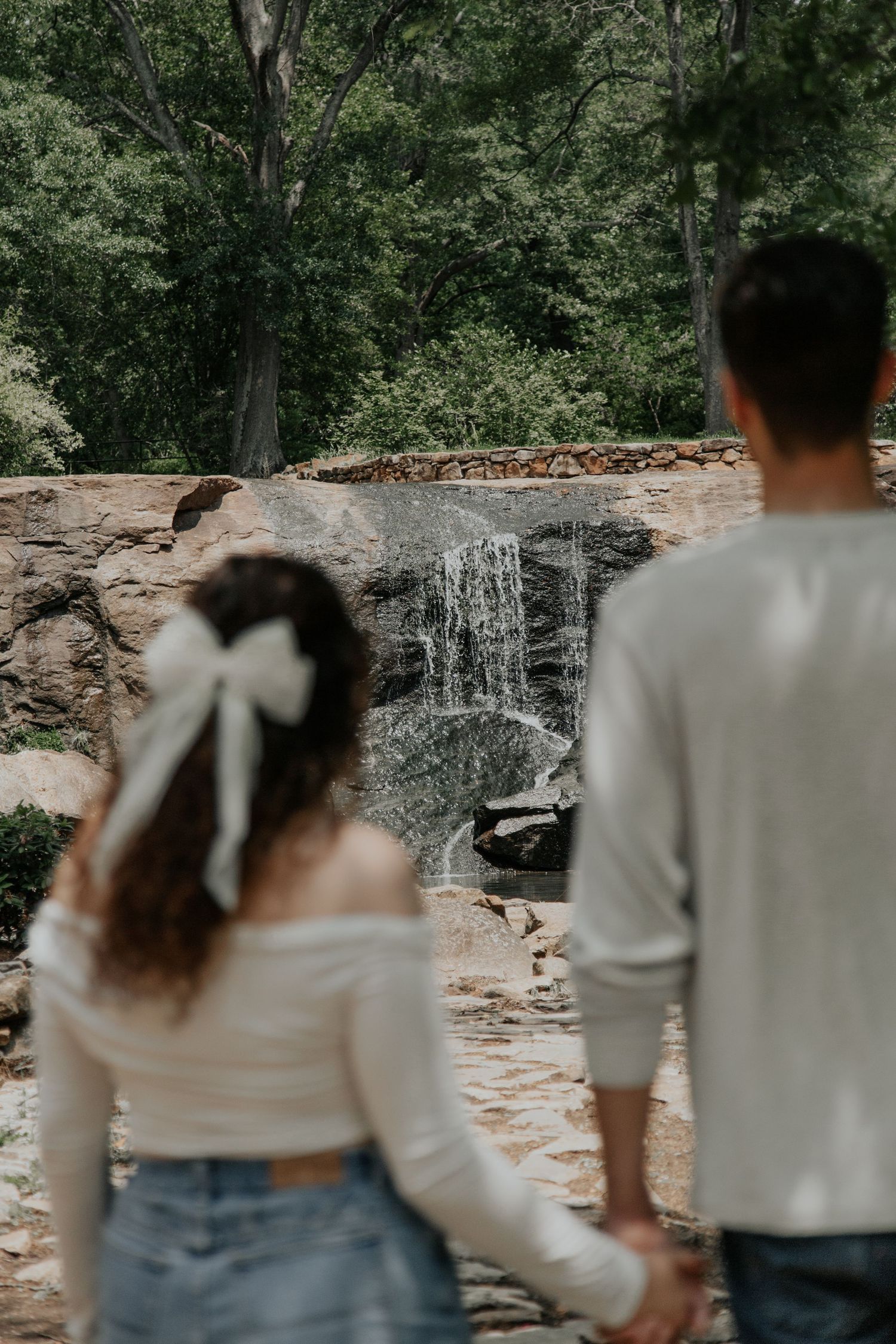 Couple holding hands facing a small waterfall surrounded by rocks and lush green trees, woman wearing white bow hair accessory.