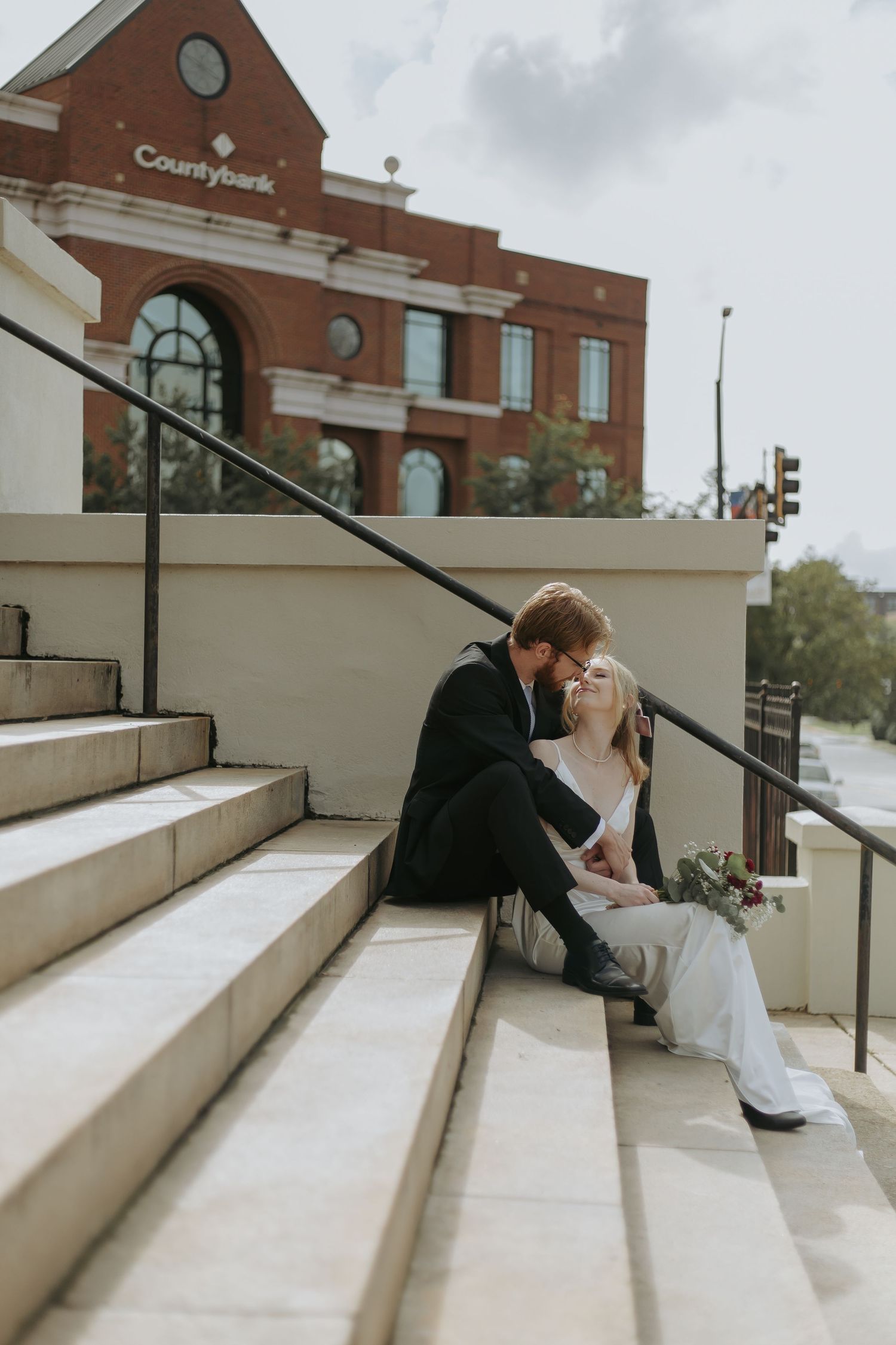 A bride and groom share a kiss while seated on outdoor steps with a brick building in the background. greenville sc