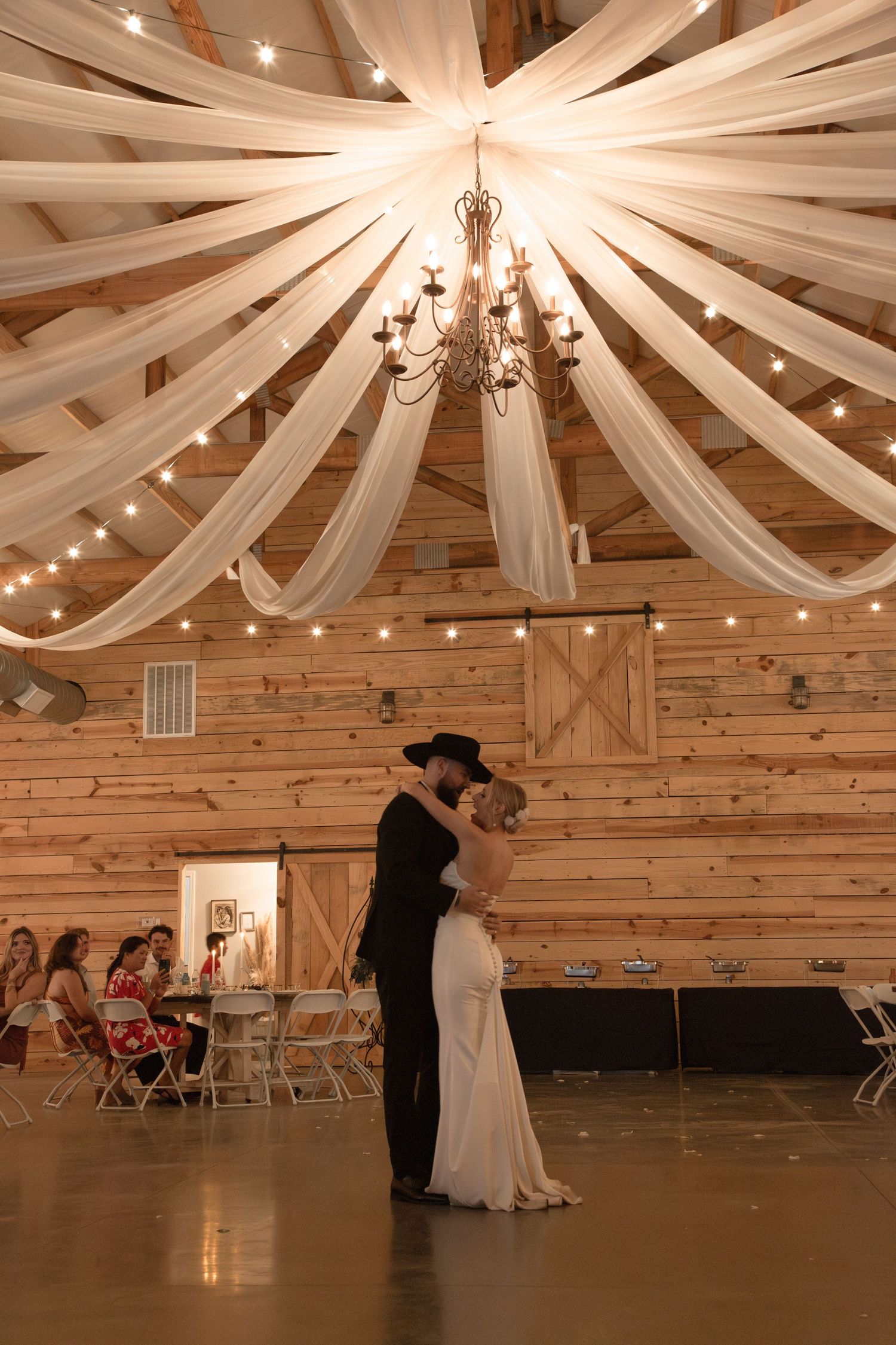 Bride and groom share romantic first dance under chandelier and draped fabric in rustic barn venue. Roger's Farm Buffalo SC.