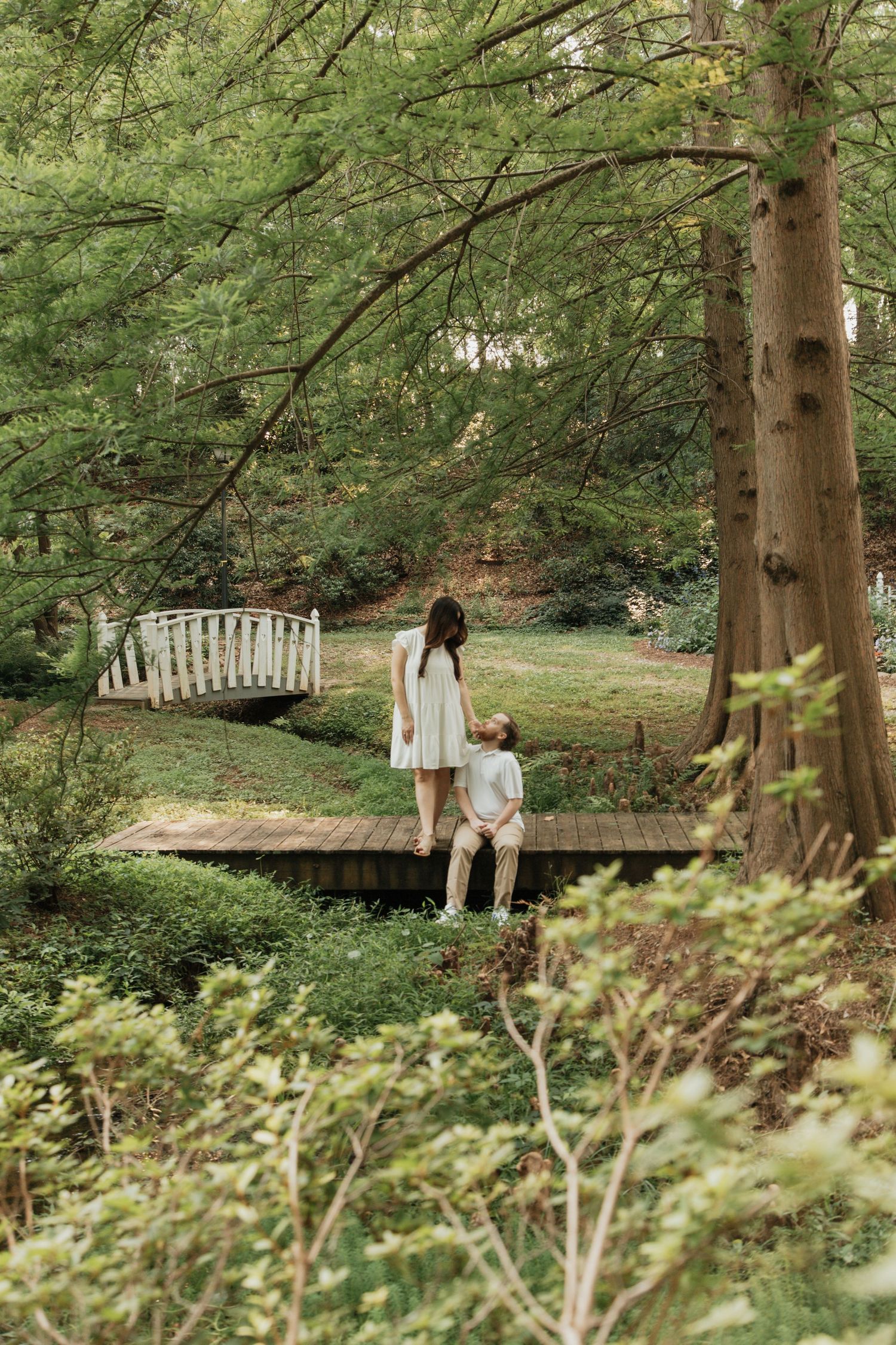 Couple standing on wooden bridge in lush green botanical garden surrounded by tall trees. kilgore lewis house and garden greenville sc