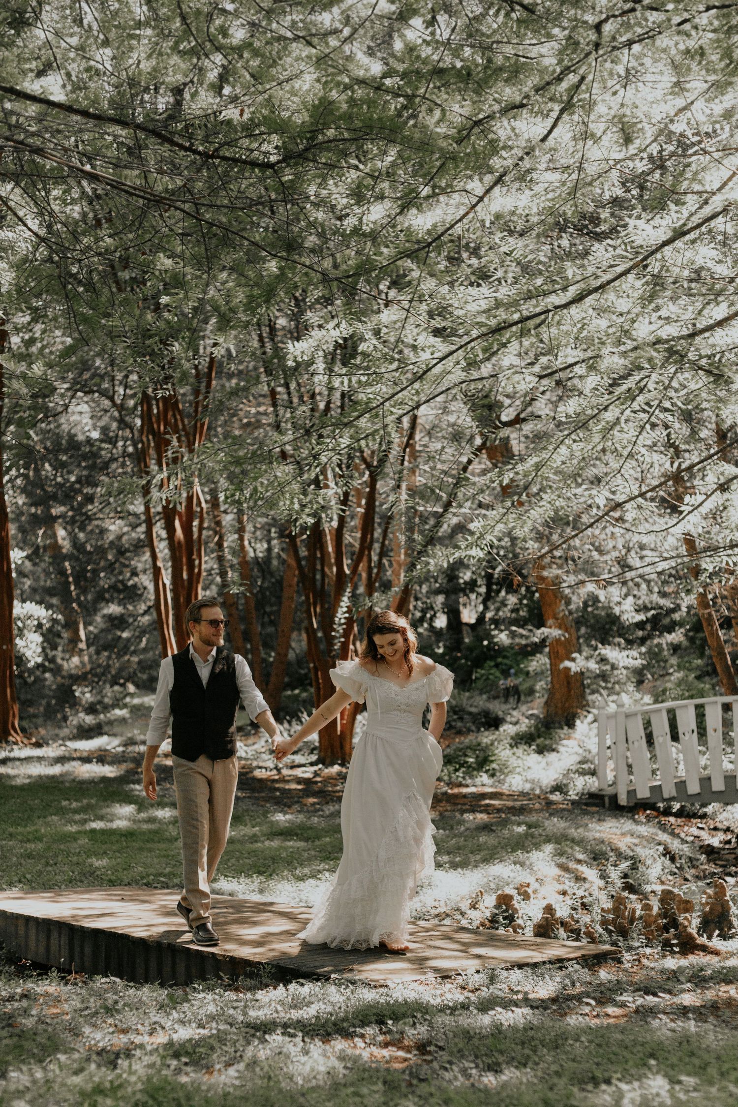 Bride and groom walking hand in hand on wooden path through sunlit forest on wedding day. kilgore lewis house and garden greenville sc