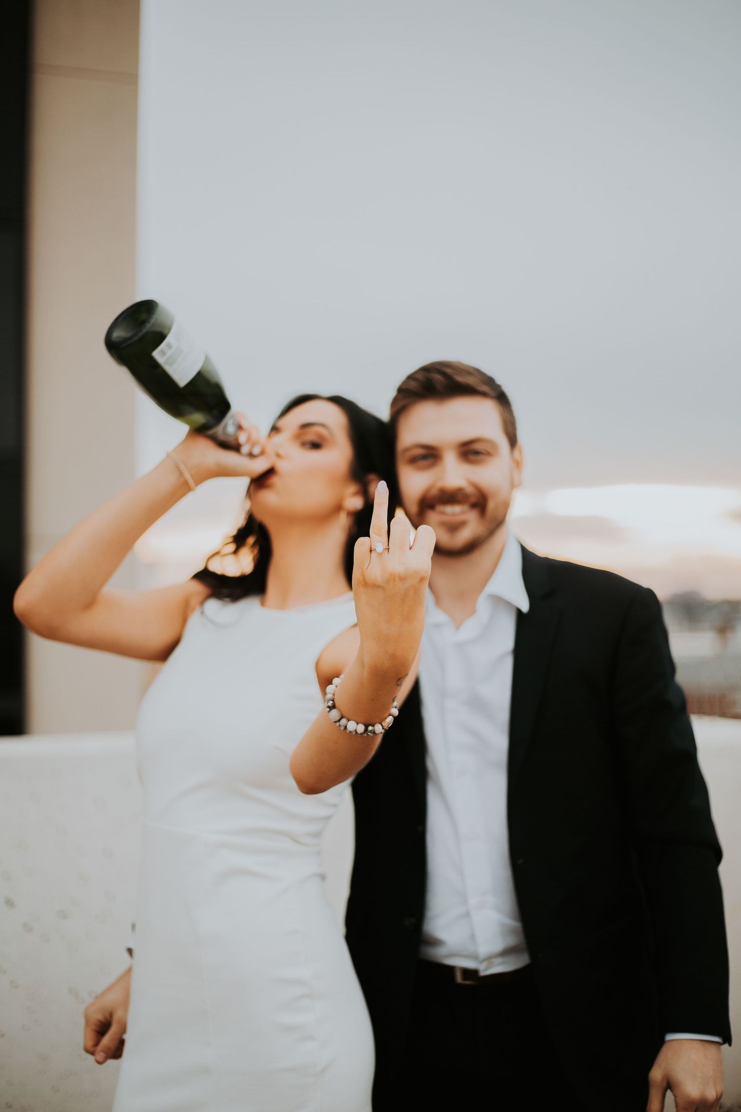 Bride in white dress drinks champagne bottle while groom in black suit smiles and gives peace sign at wedding.
