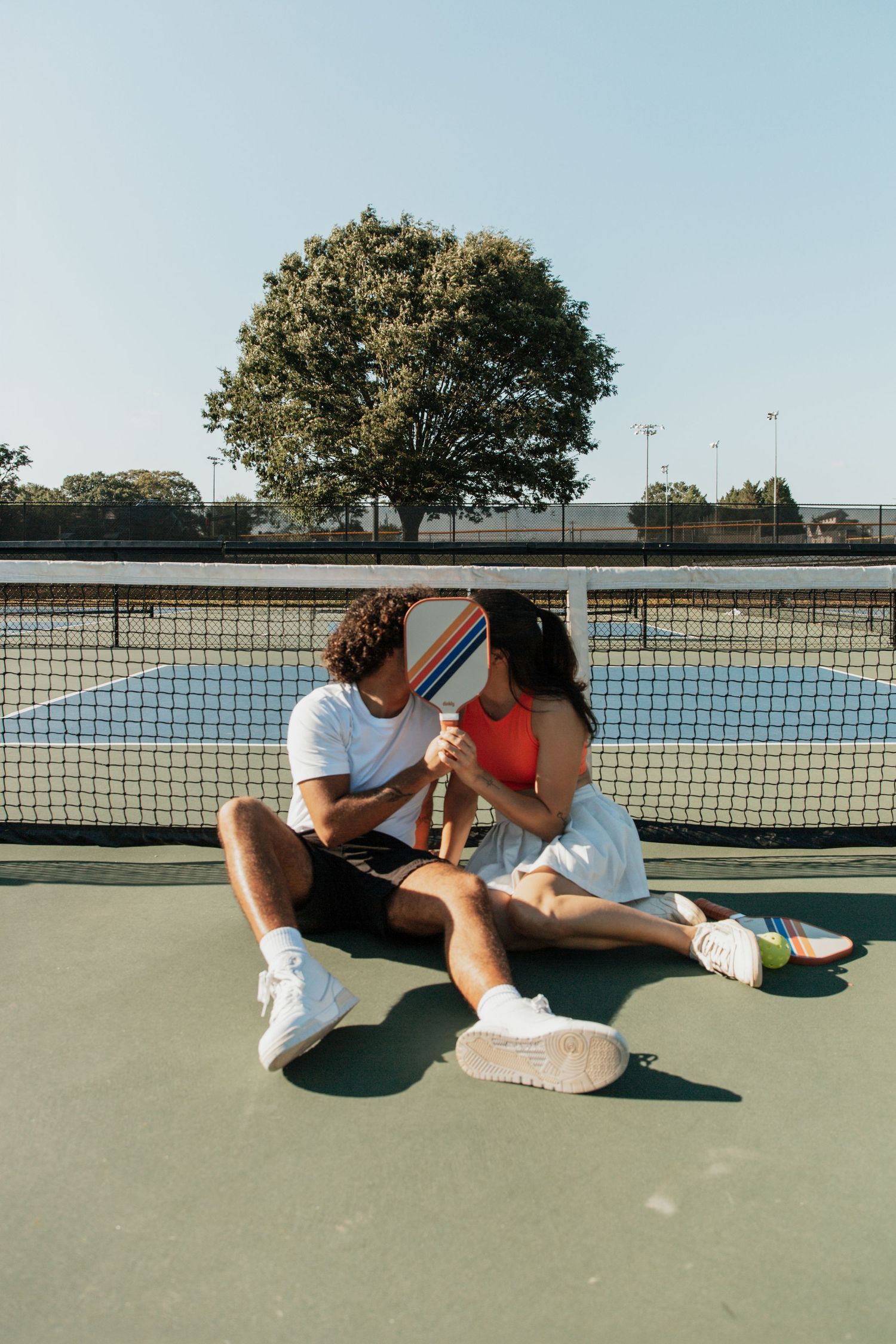 Couple kissing behind a pickleball paddle while sitting on an outdoor court near the net on a sunny day.