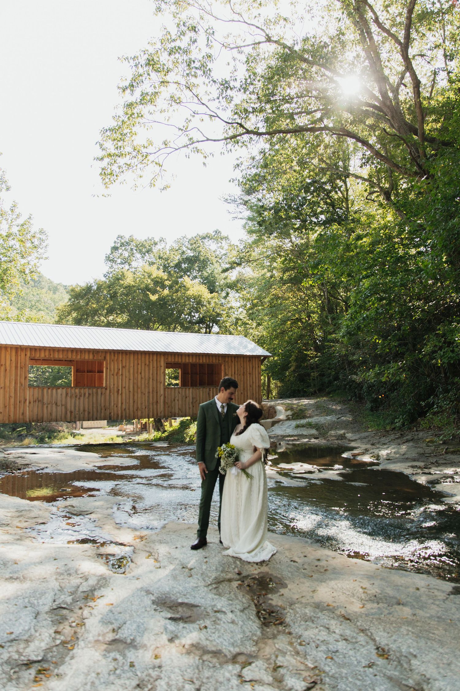 Bride and groom standing on rocky creek bed before rustic covered bridge surrounded by lush forest. venue at ballenger bridge landrum sc