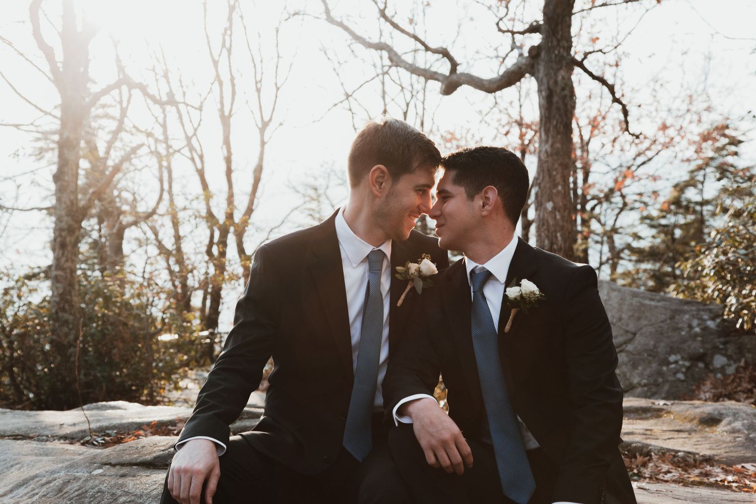 Two grooms in dark suits share an intimate moment seated outdoors at their wedding ceremony.