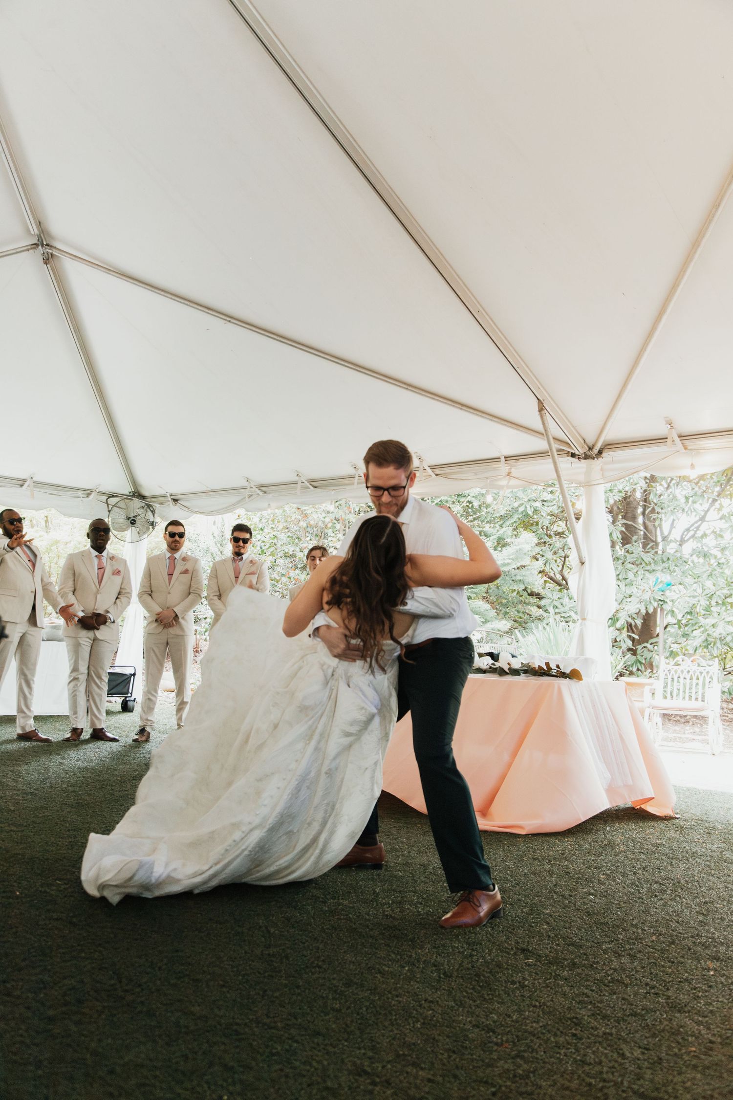 Bride and groom share first dance under white tent at outdoor wedding reception with groomsmen watching. Viewpoint at Buckhorn Creek.