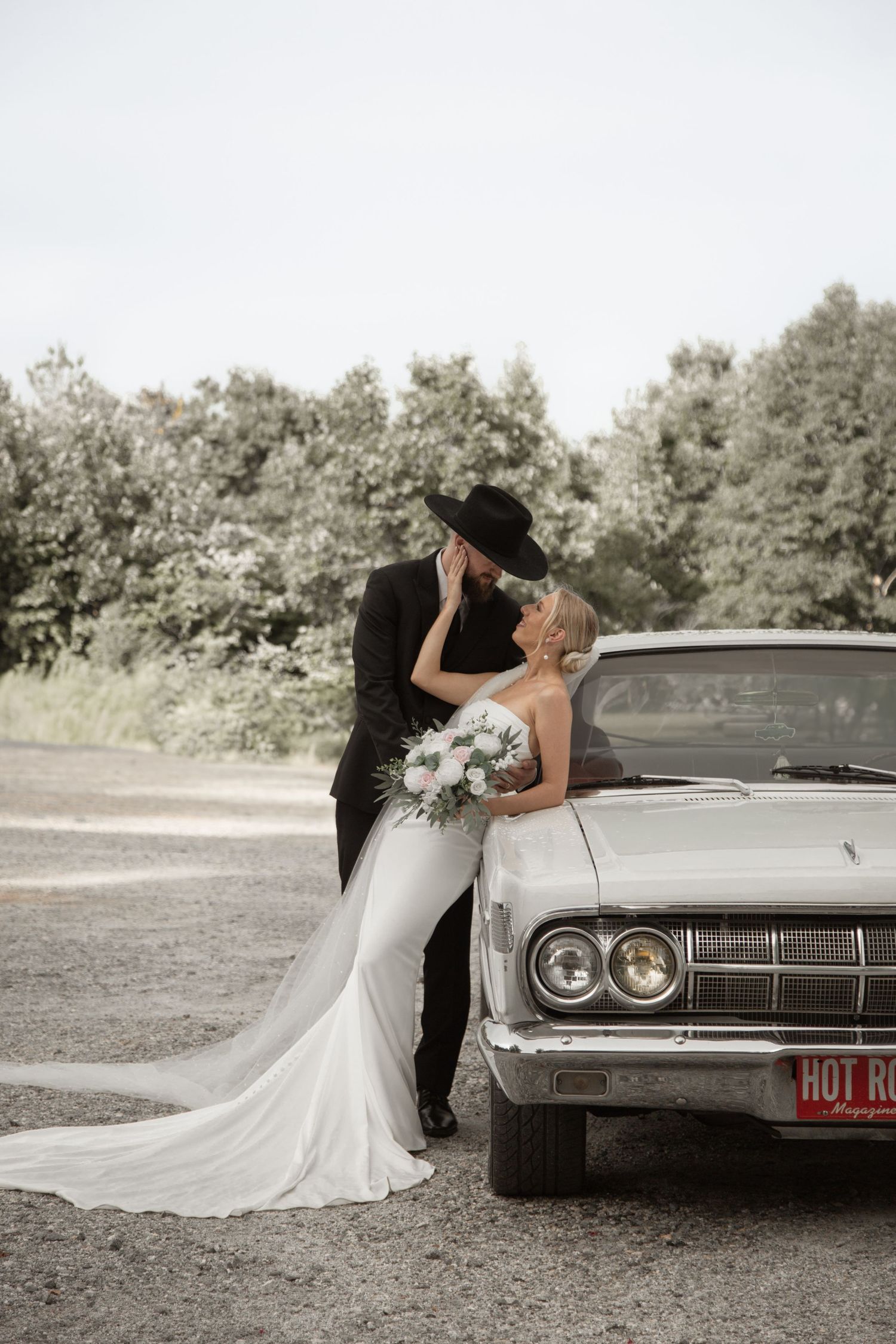 A bride and groom kiss beside a vintage white convertible on a gravel road surrounded by lush trees. rogers farm buffalo sc