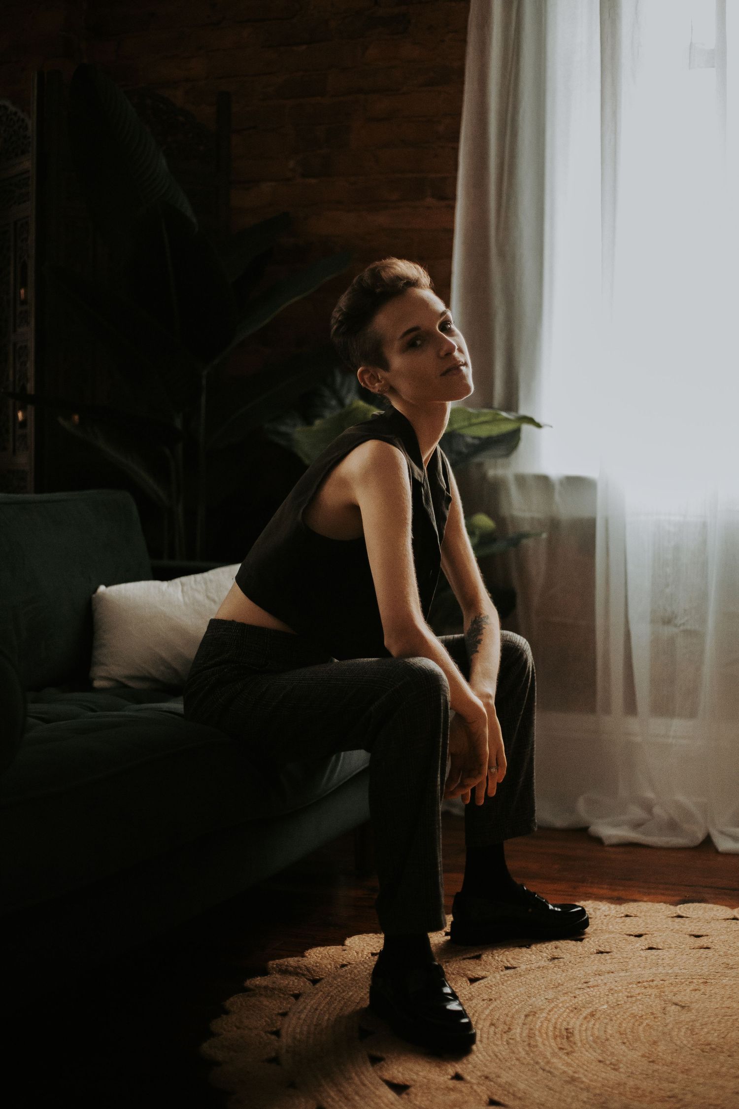 Young woman sitting pensively on green sofa near bright window in moody dark interior with tropical plants. wanderlust boudoir studio