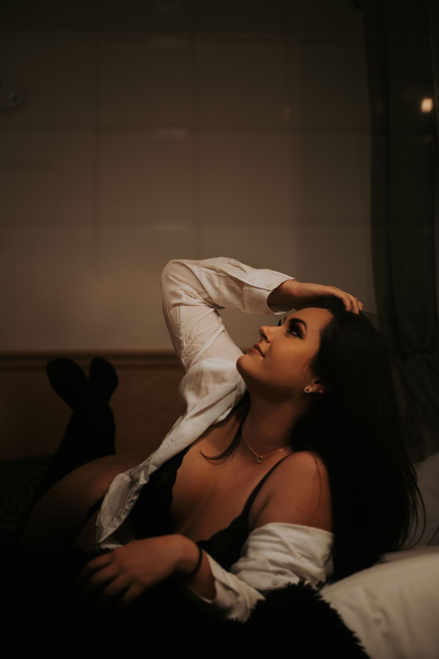 Woman in black lingerie and open white shirt posing on a bed in dim warm lighting.