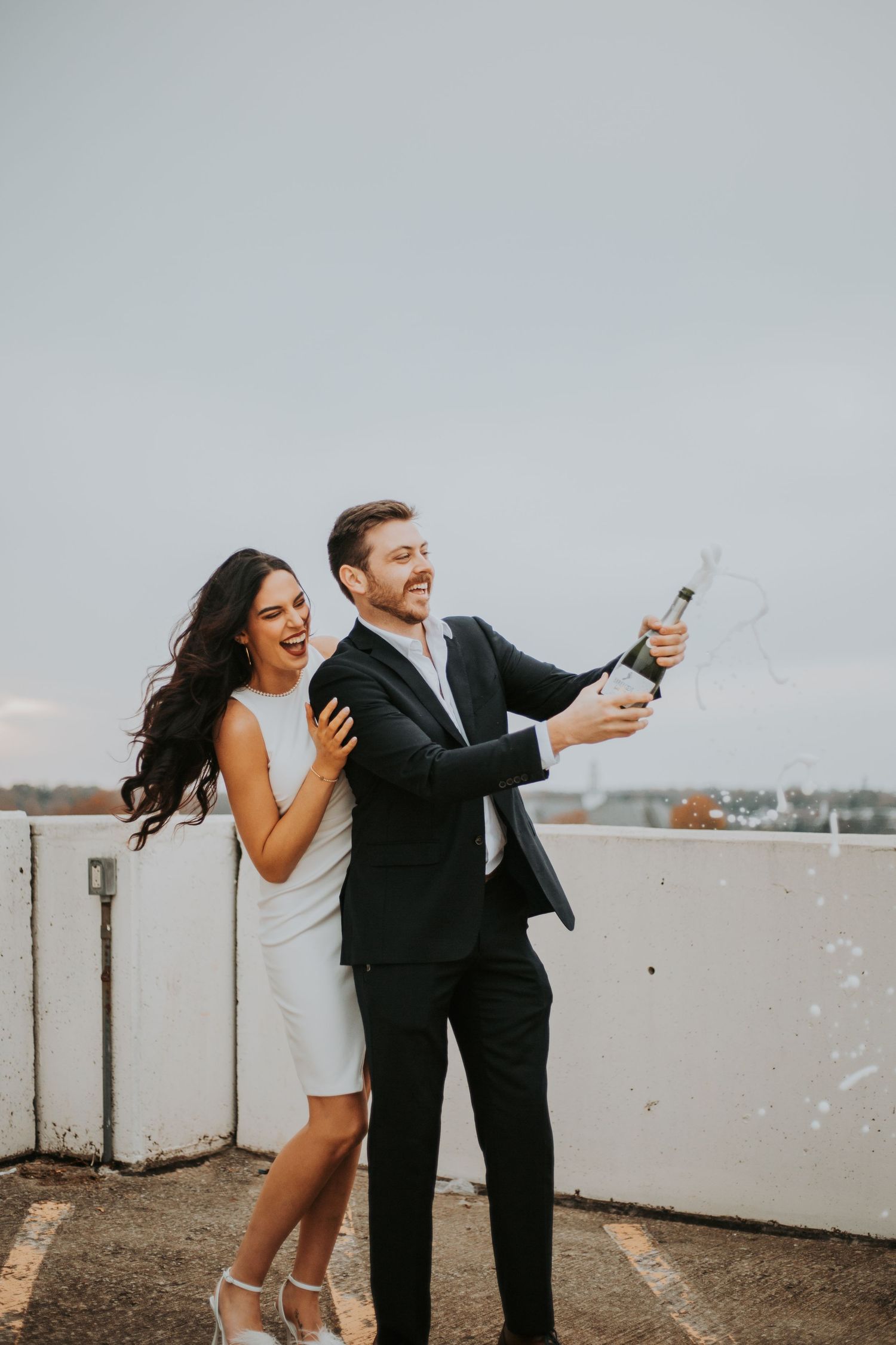 Joyful couple popping champagne on a rooftop, woman in white dress and man in black suit. greenville sc